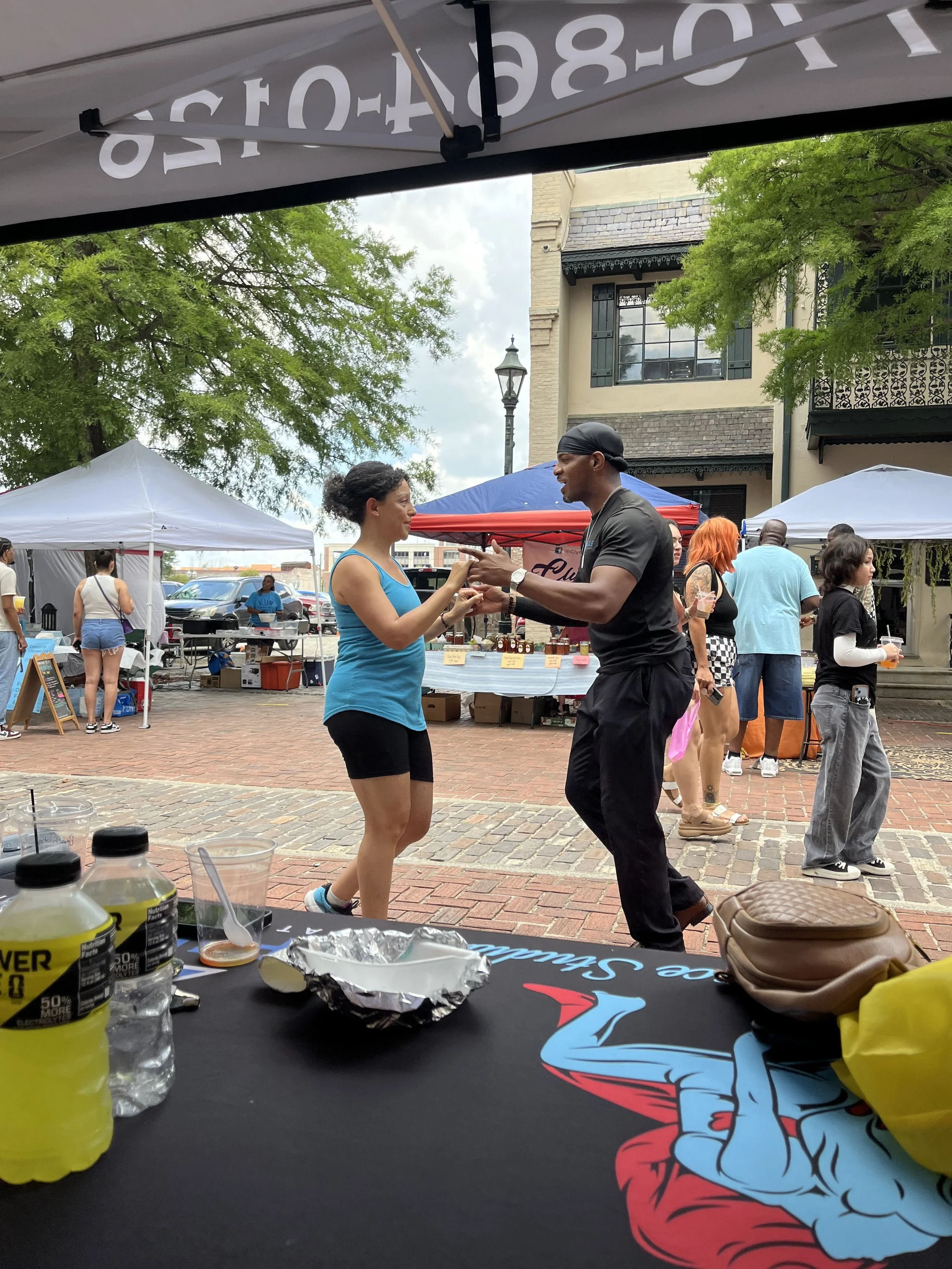 People dancing and socializing at an outdoor event with tents, trees, and buildings in the background. The foreground shows bottles, a foil-wrapped food item, and a black cloth with a colorful graphic design.