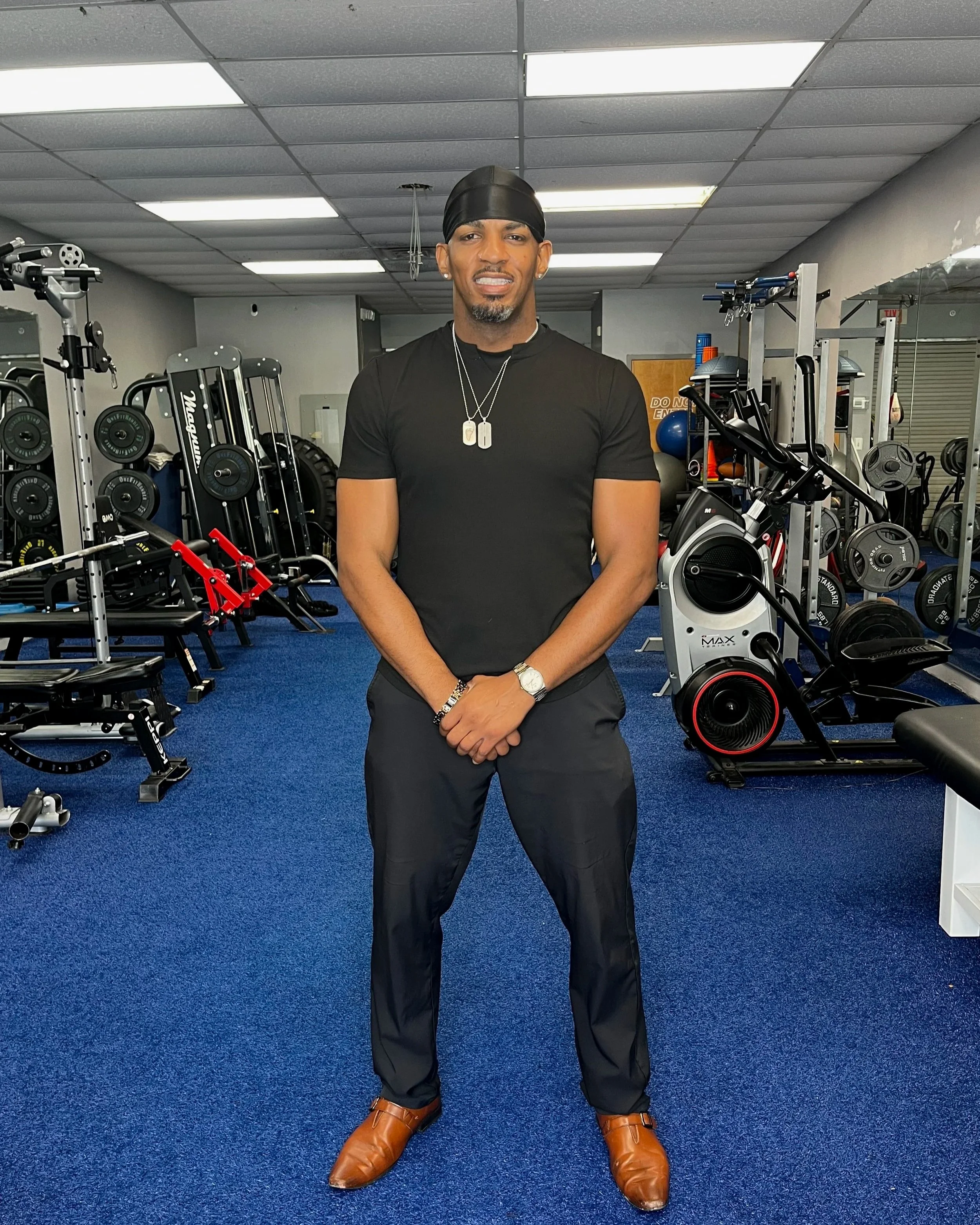 A man standing in a gym, wearing a black t-shirt, black pants, brown boots, silver necklaces, and a black headscarf, with various fitness equipment around him.