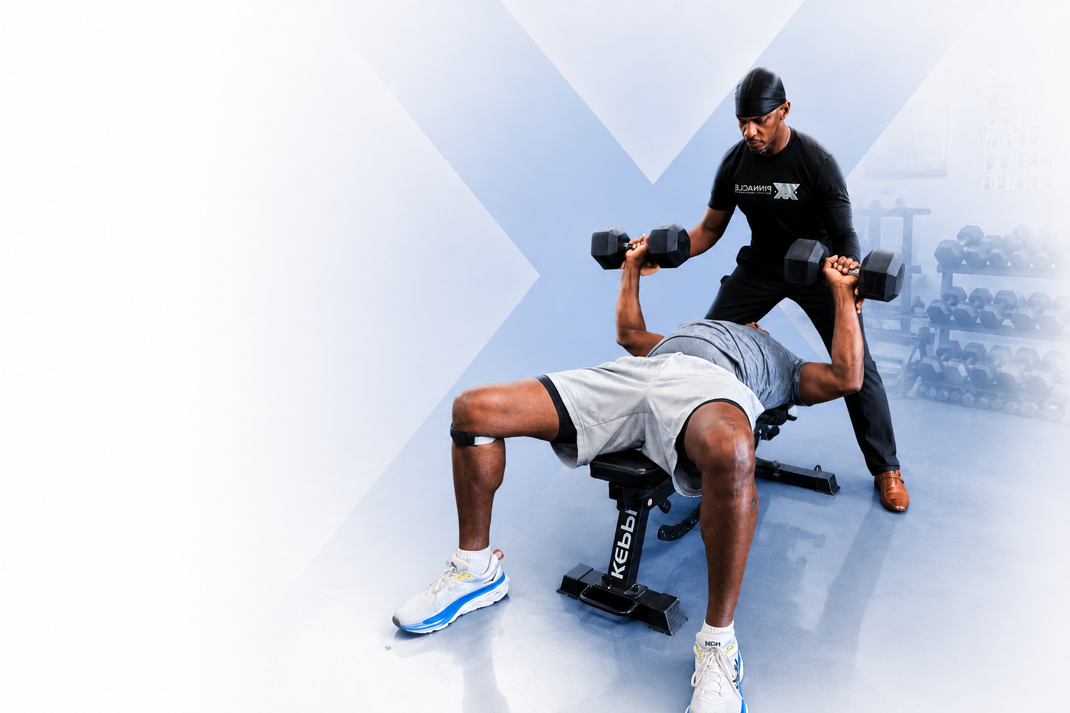 Two men in a gym, one lying on a bench press, the other spotting him with dumbbells.