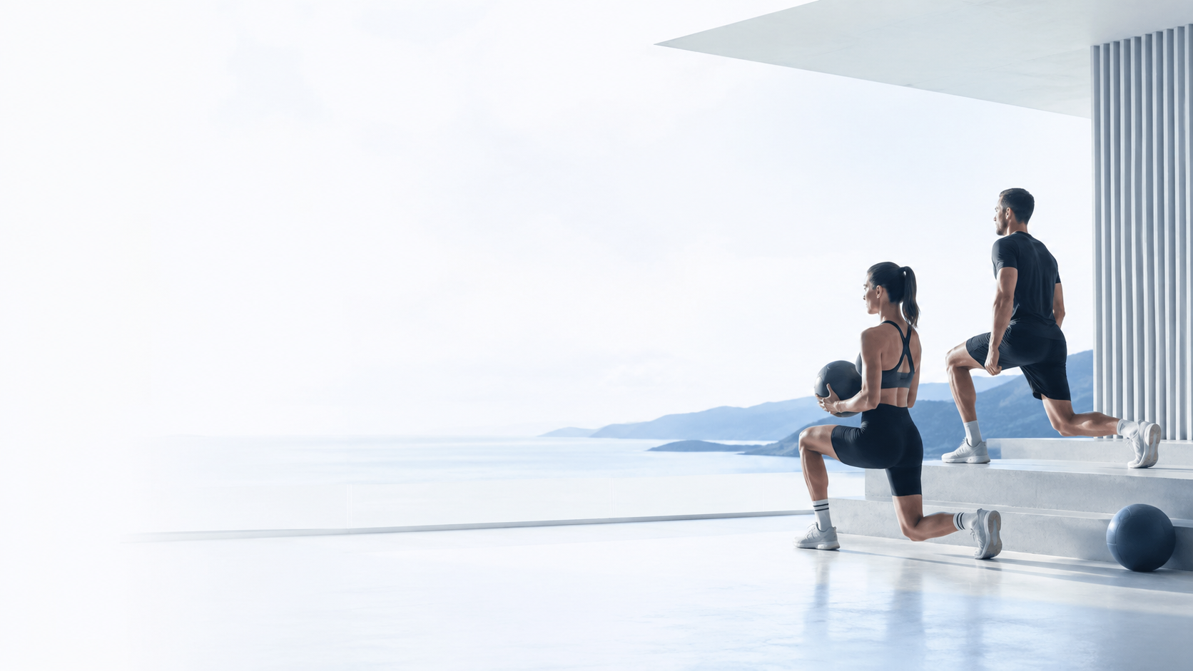 Two people exercising on stairs with a scenic view of mountains and water in the background. The woman is holding a medicine ball, and the man is lunging upwards.