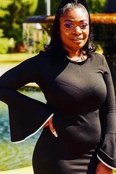 A woman in a black dress with bell sleeves standing outdoors by a pond or lake.
