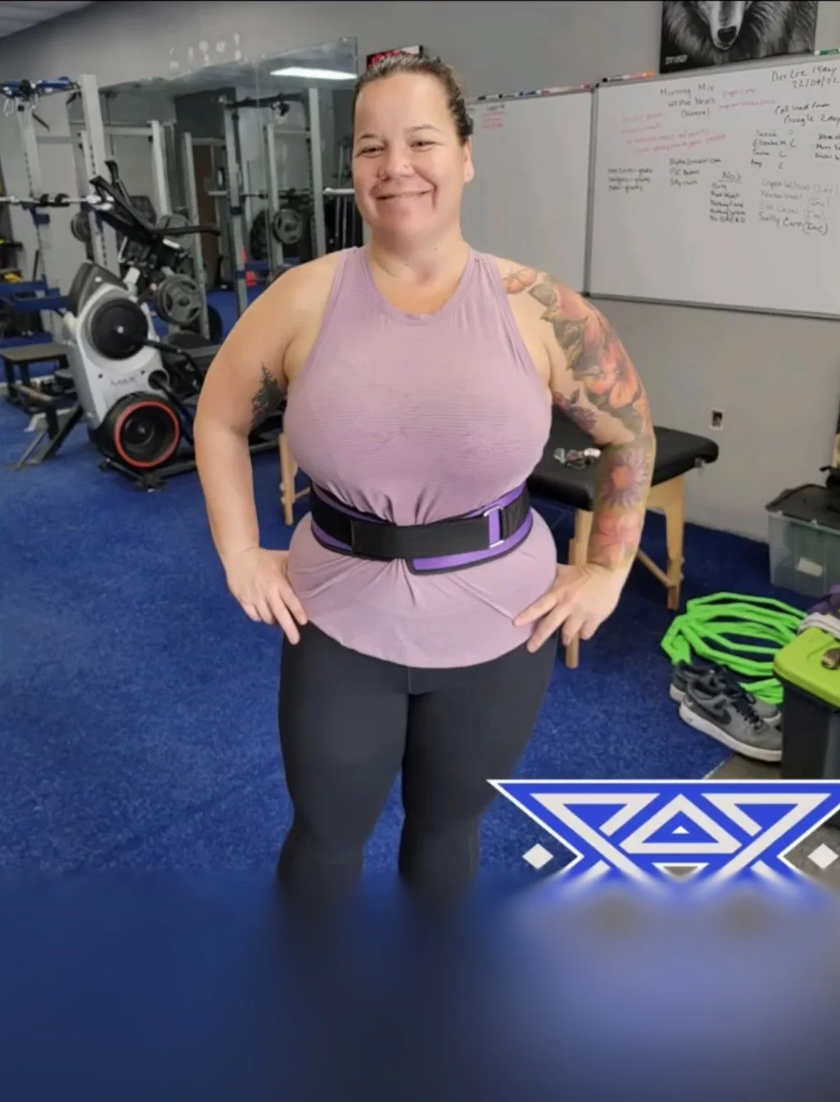 A woman smiling in a gym, wearing a light purple athletic top, black leggings, and a black and purple belt. In the background, there is gym equipment, a whiteboard with notes, and various fitness accessories on the floor.