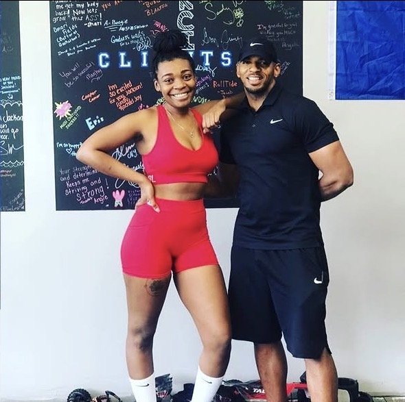 A woman in a red sports bra and shorts smiling with her arm around a man in a black Nike athletic outfit, standing in front of a blackboard with handwritten messages.