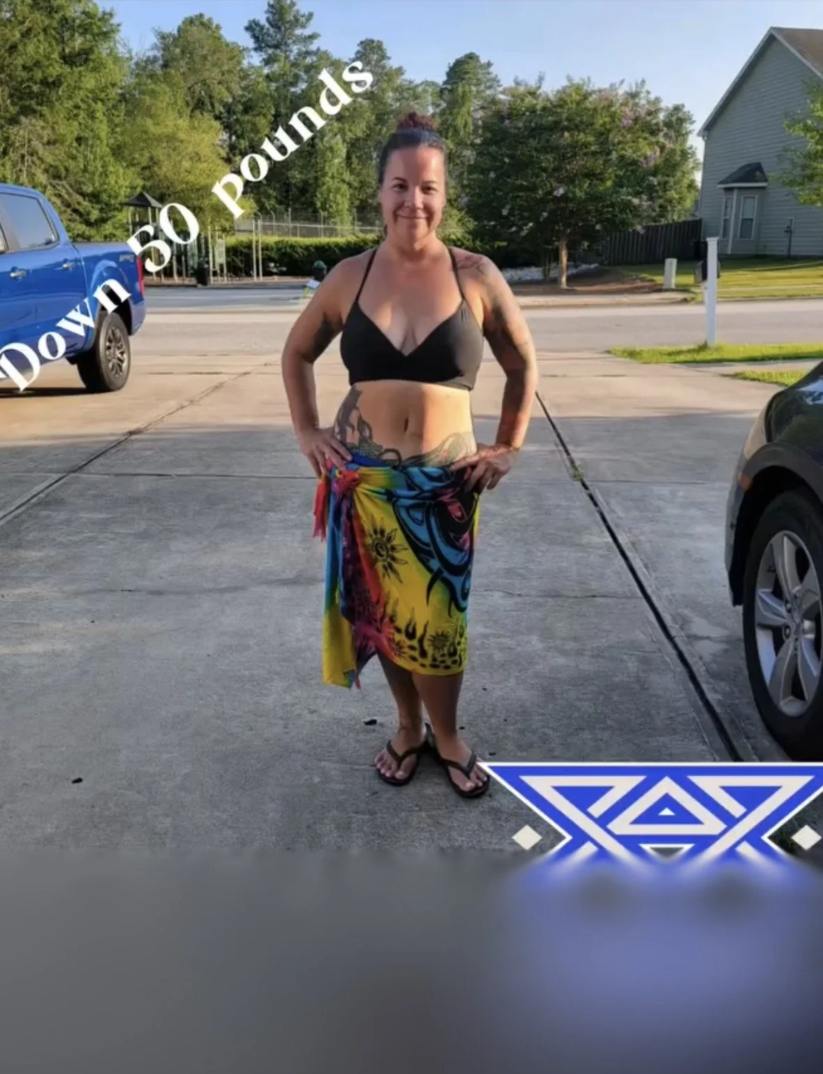 A woman stands outside on a driveway, smiling, wearing a black bikini top and a colorful tropical skirt. She has tattoos on her arms and waist, and her hair is tied back. In the background, there are trees, a house, and a parked blue truck.