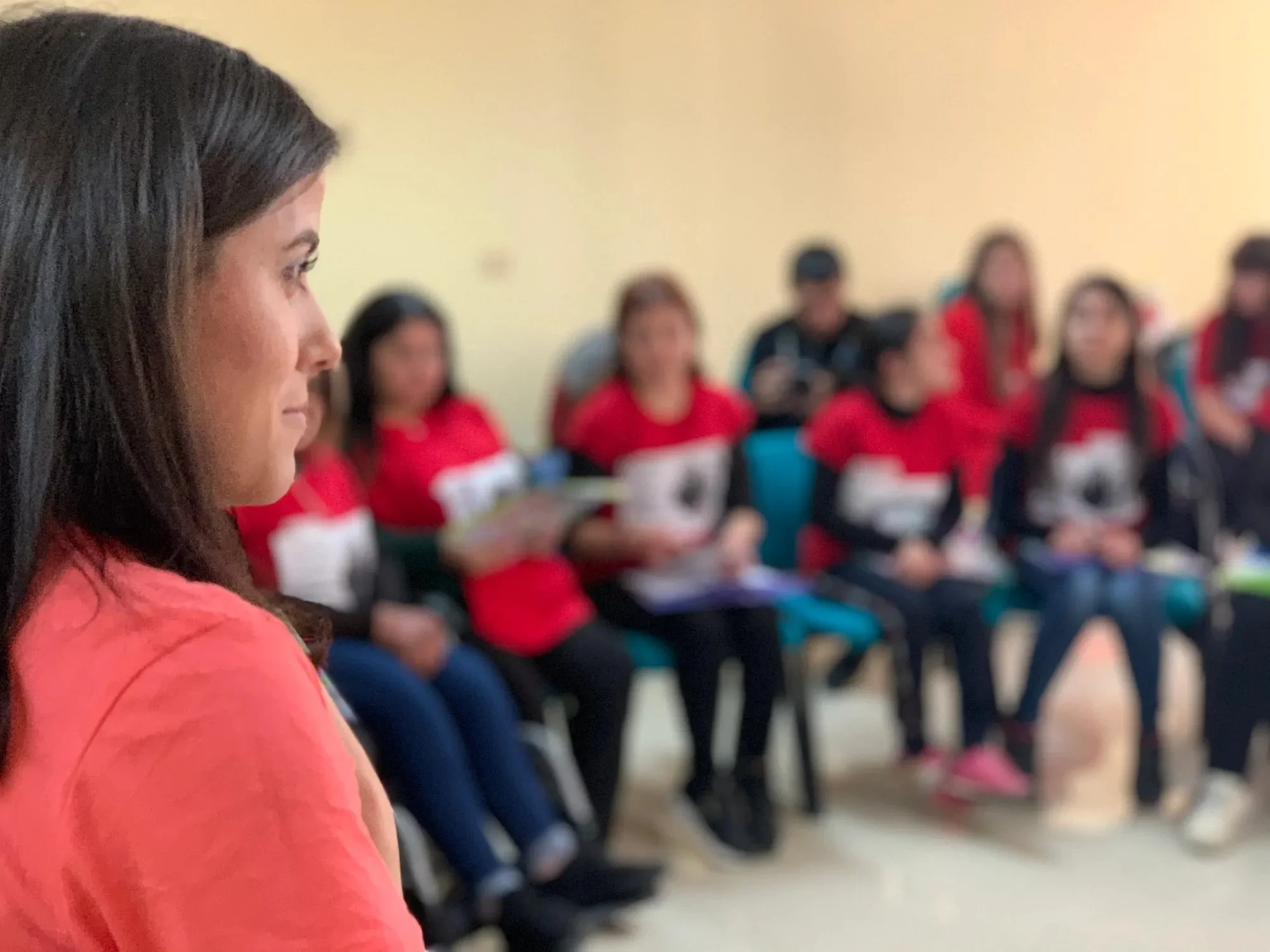 A woman with dark hair in a coral top is sitting in profile, facing a group of people in a classroom or meeting room.