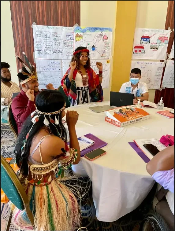 A group of people at a round table during a cultural presentation or discussion, with posters on the wall behind them, some wearing traditional or costume accessories, and one person speaking or presenting.
