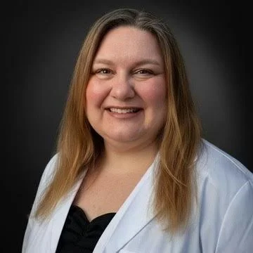 A woman with long light brown hair wearing a black top and white lab coat, smiling against a dark background.