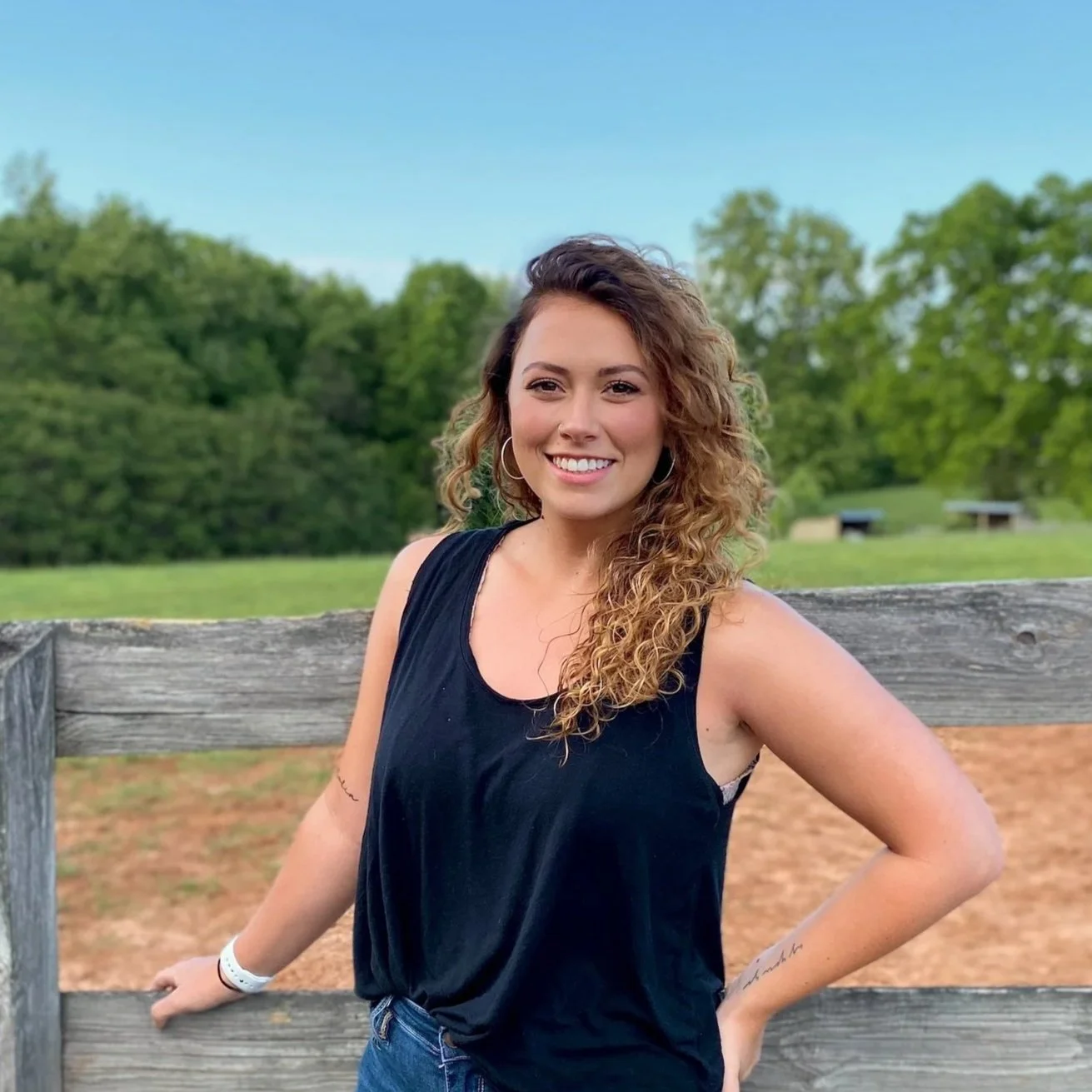 A young woman with curly hair, wearing a black sleeveless top, smiling and standing outdoors with a wooden fence and green trees in the background.