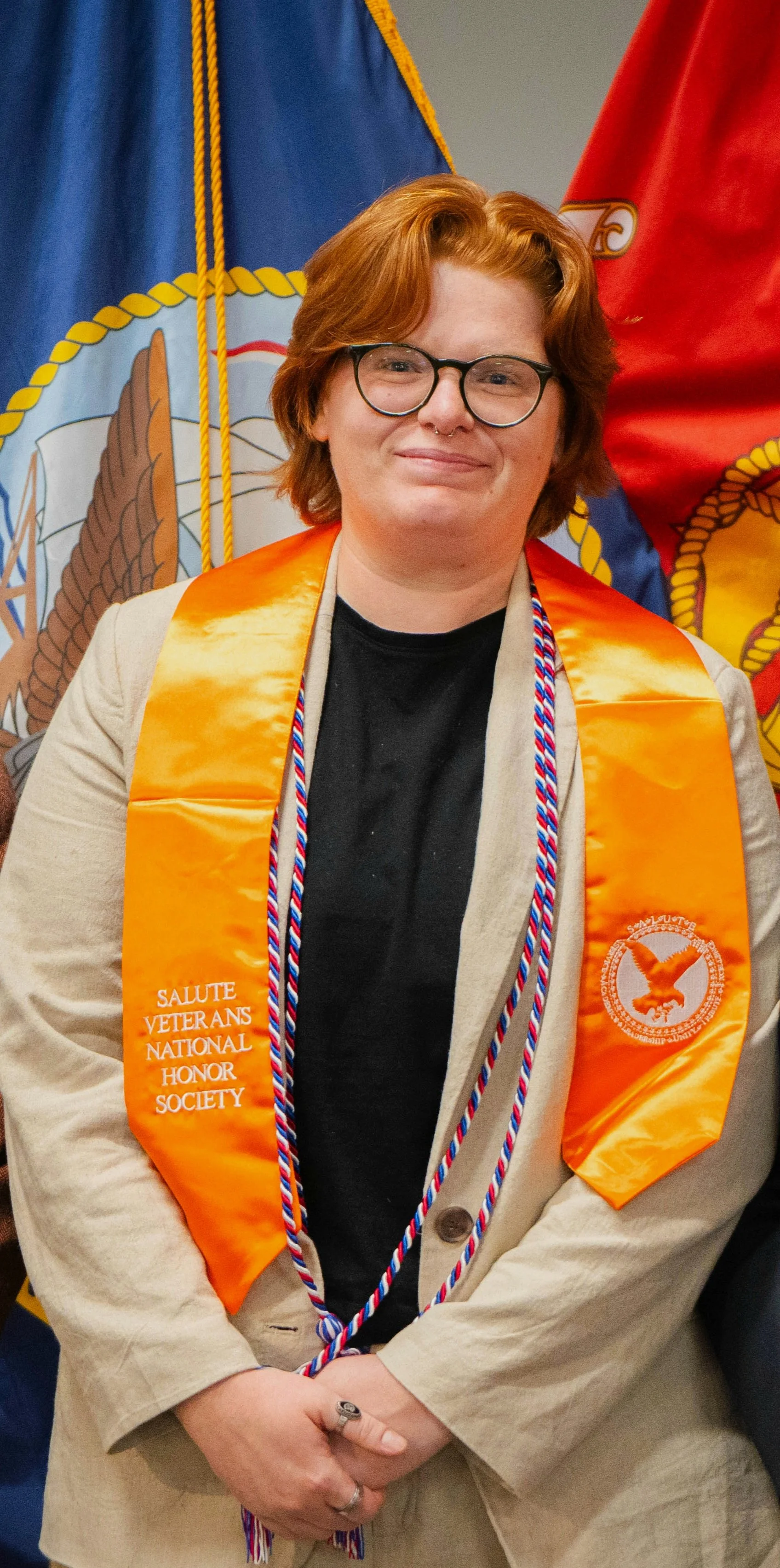 A smiling person with short red hair wearing glasses, a beige blazer, and an orange sash that reads 'Salute Veterans National Honor Society' stands in front of a blue and red banner.