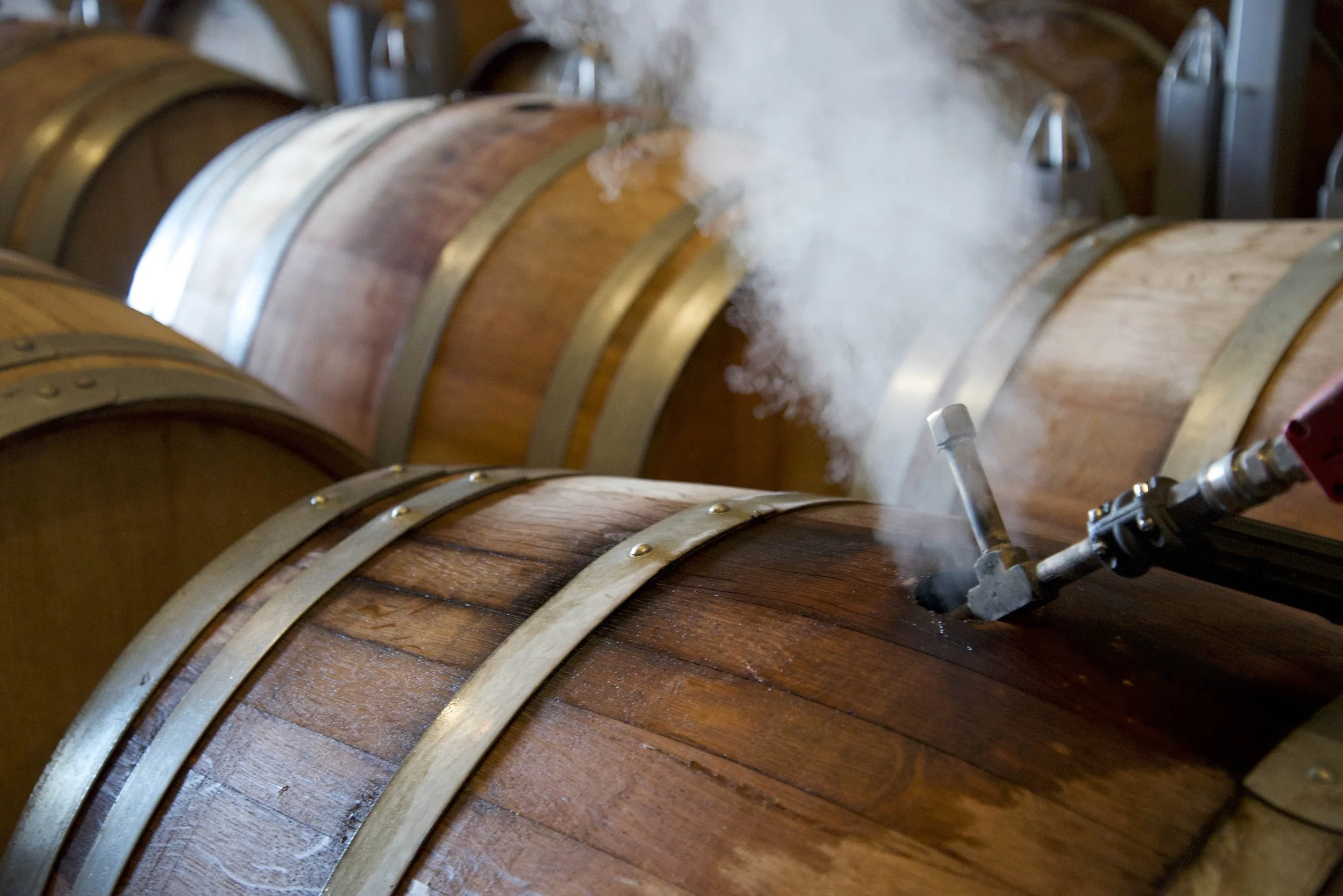 A worker a winery using a tool that's steaming into a wooden wine barrel.