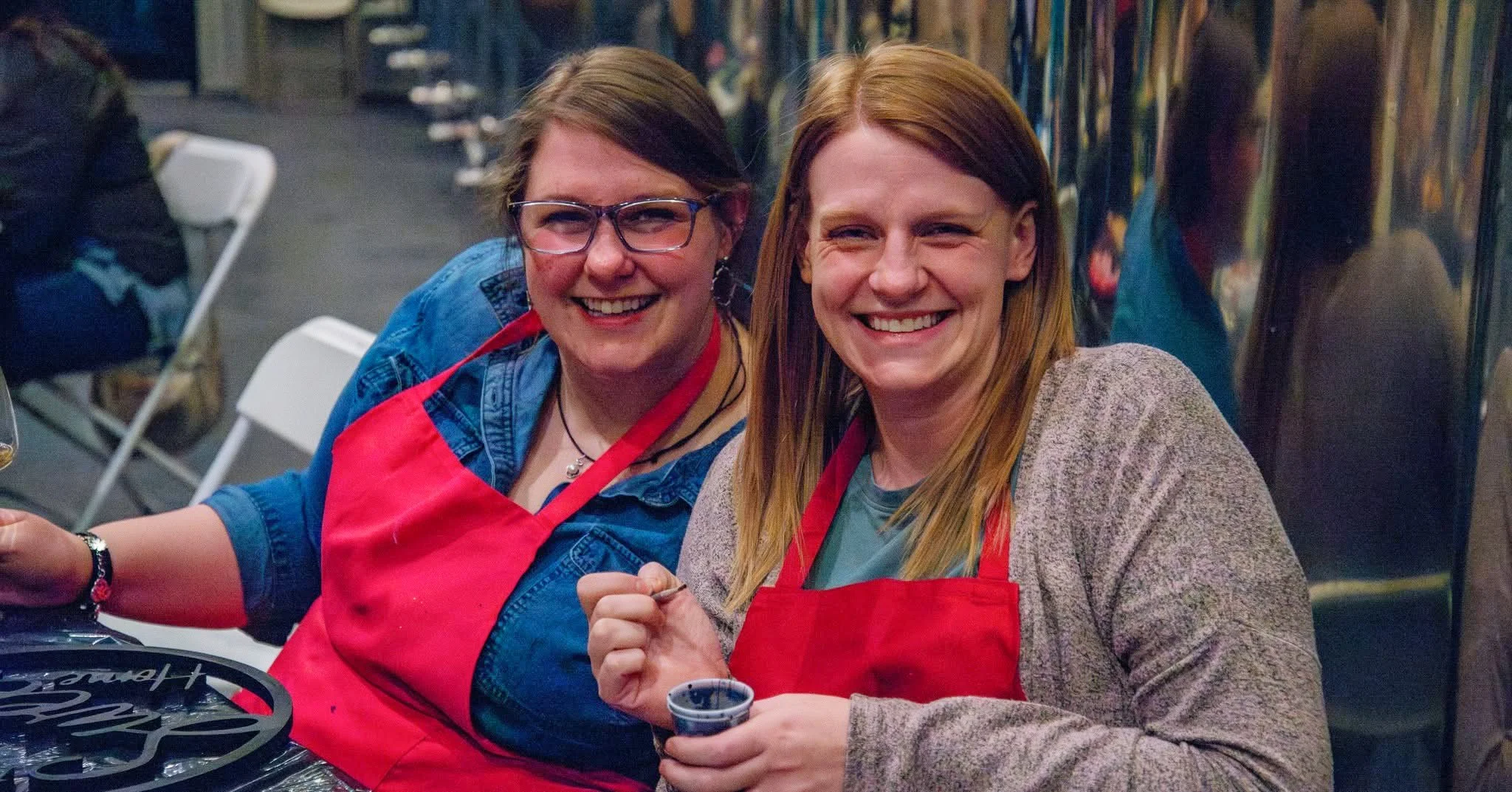 Two women smiling and sitting side by side at a table, wearing red aprons, at a Bar Z Winery event.