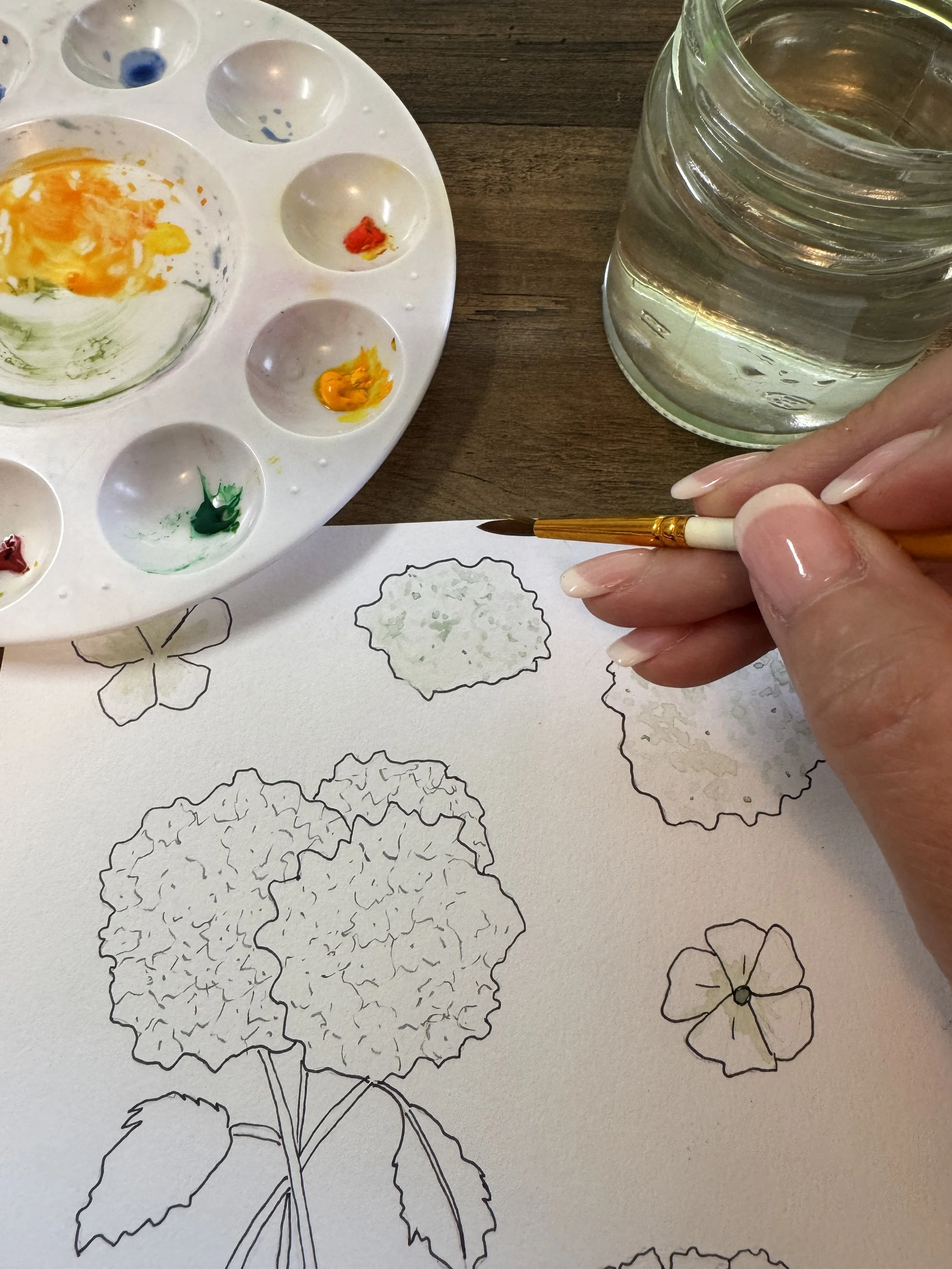 An artist hand holding a small paintbrush near a glass jar of water. An artist's palette with colorful paint blobs and a partially painted floral sketch outline on paper on a desk surface.