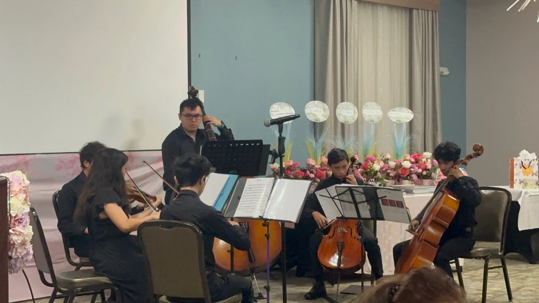 A musical performance featuring a group of young musicians playing string instruments, including violin and cello, in front of an audience. The performers are dressed in black, and there are floral decorations and balloons in the background.