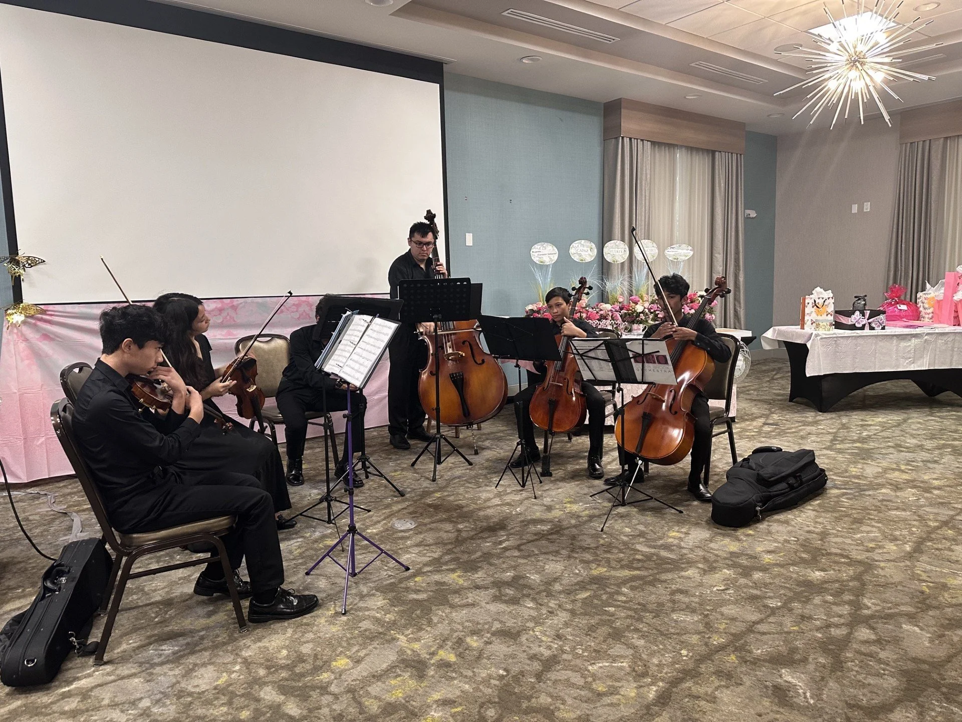 A group of young musicians playing string instruments, including violins and cellos, during a performance in a decorated banquet hall with floral arrangements and gift bags on a table in the background.