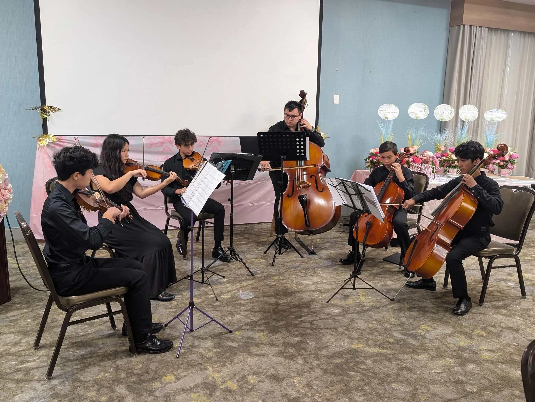 A group of young musicians performing in a room decorated with pink flowers and floral-themed balloons. The musicians are playing string instruments, including violins and a double bass.