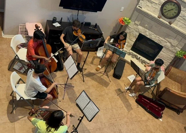 A group of six young people playing string instruments in a living room, sitting on chairs arranged in a semi-circle, with music stands in front of them. The room has a fireplace, a TV mounted on the wall, a large clock, and some decorative items including potted plants and flowers.
