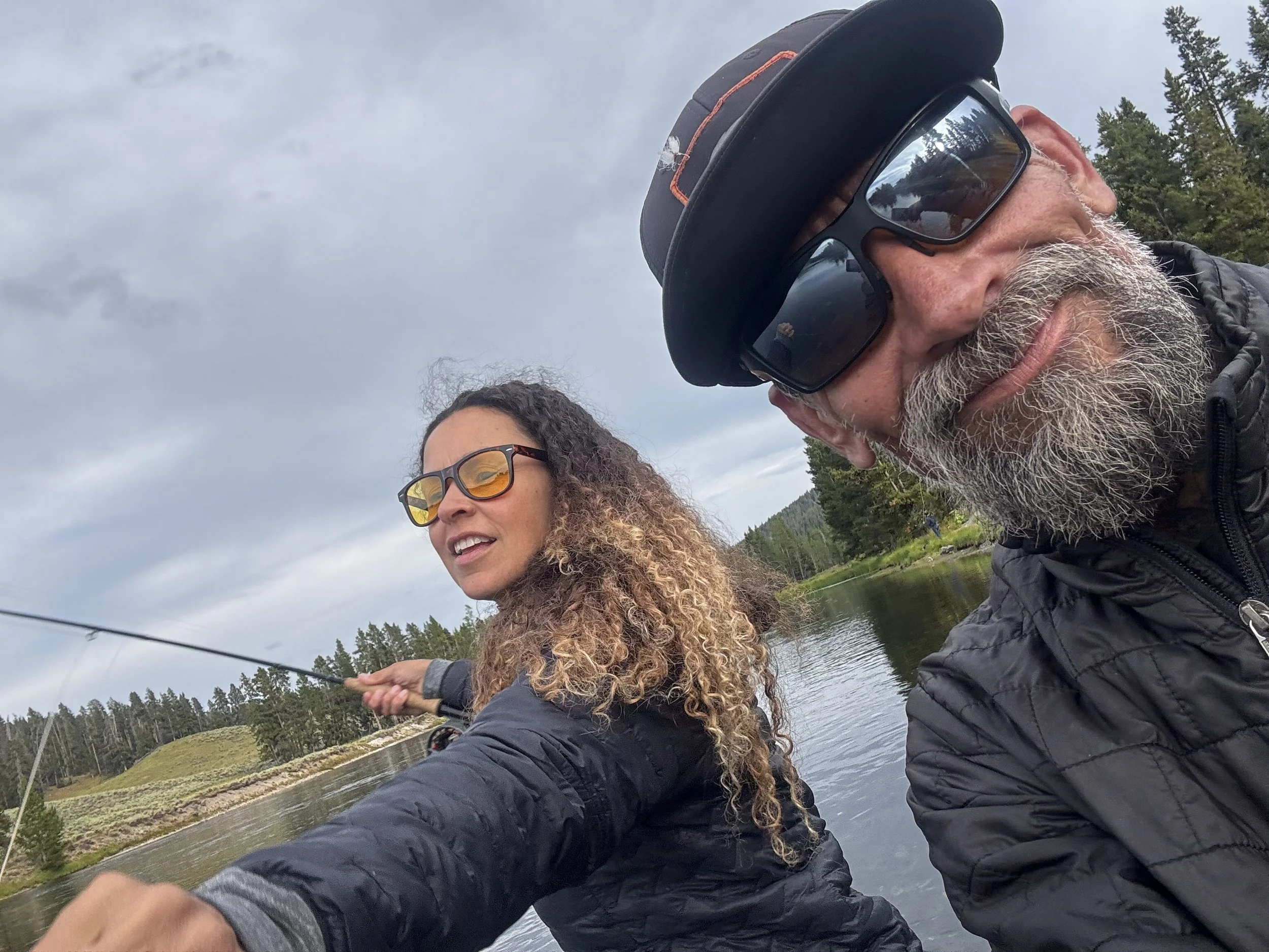 Two people, a man and a woman, are fly fishing by a river in an outdoor setting with trees and mountains in the background. They are wearing glasses and jackets, and the man is wearing a hat and sunglasses.