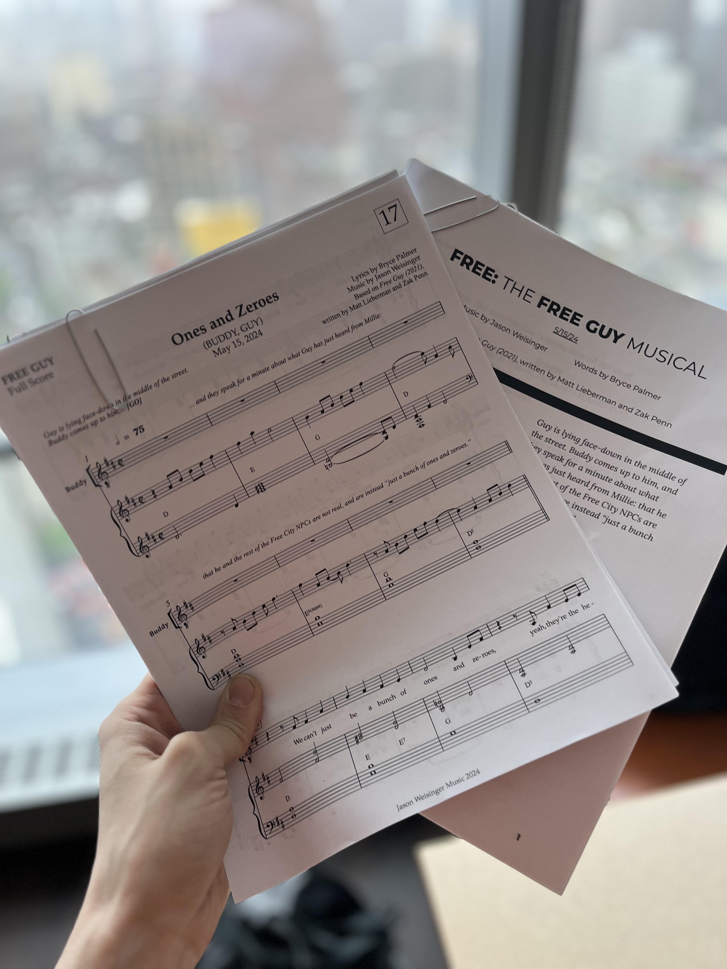 Hand holding sheet music with song title 'Ones and Zeroes' by Buddy Guy, and another sheet titled 'FREE: THE FREE GUY MUSICAL' with music credits, against a blurred cityscape background.