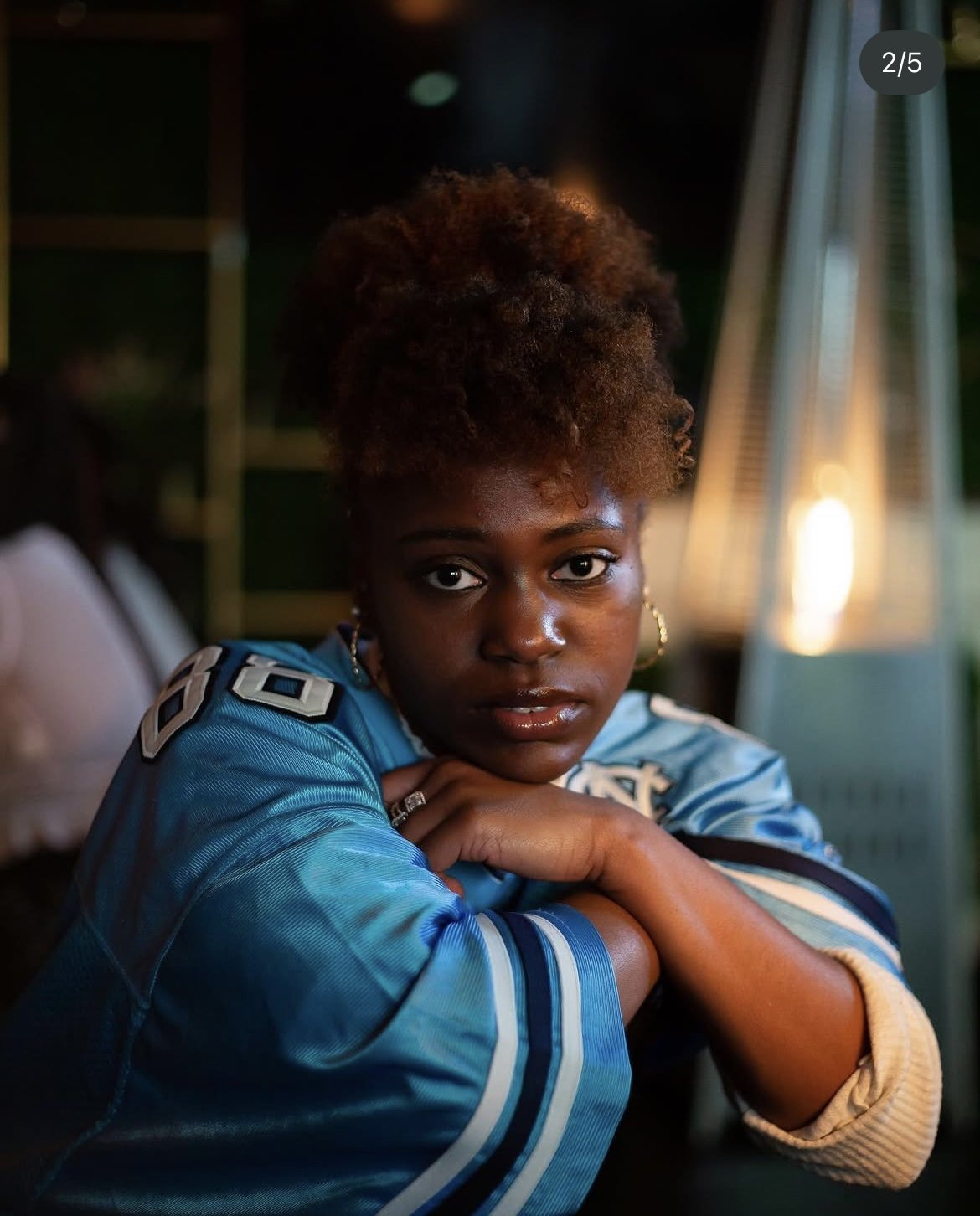 Portrait of a young woman with natural curly hair, wearing a blue sports jersey, posing indoors with her arms crossed on a table.