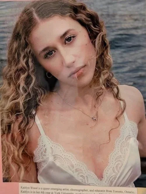 A young woman with curly blonde hair and light skin is looking directly at the camera. She is wearing a white lace-trimmed top and a delicate necklace with a letter 'K'. The background appears to be a body of water.