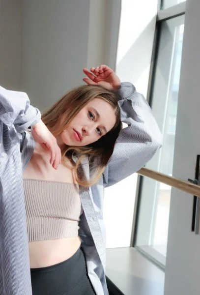 Young woman with shoulder-length brown hair and light skin, posing indoors near a large window, wearing a light beige crop top and a gray unbuttoned shirt.