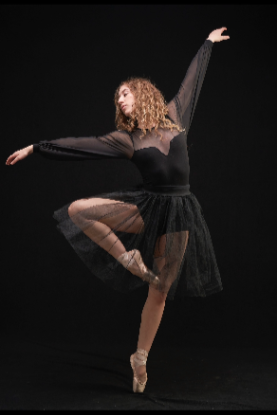 A ballet dancer in a black costume performing a pose on pointe with one leg raised and arms extended.