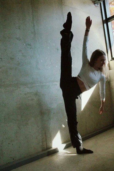 A woman in a white long-sleeve shirt and black pants is balancing on one hand against a concrete wall while doing a high leg stretch with her other leg up and her hand reaching toward the ceiling.