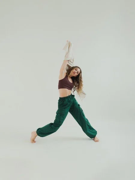 A young woman with curly hair dancing or posing in a studio with a plain, light-colored background. She is wearing a dark tank top and loose green pants, holding a white cloth in one hand, with her other hand raised above her head.