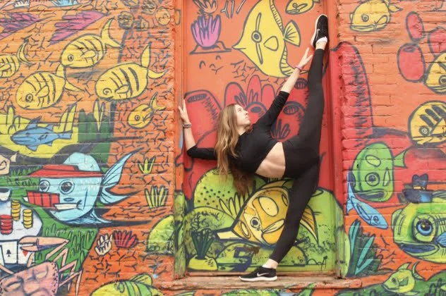 A woman in black athletic wear is performing a standing split pose against a colorful graffiti mural of various aquatic creatures, including fish, in an outdoor setting.