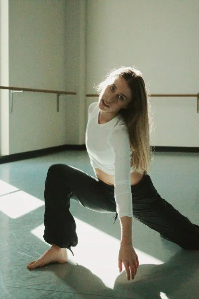 A woman with long hair wearing a white top and black pants is stretching in a dance studio with a wooden ballet barre along the wall.