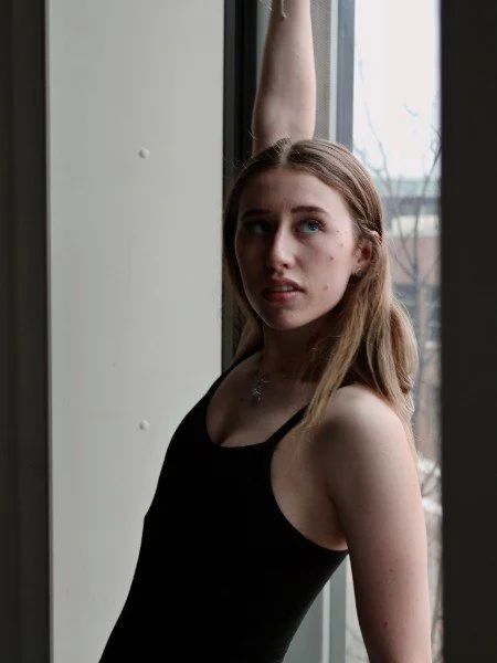 Young woman in black tank top standing by window with one arm raised, looking at camera.