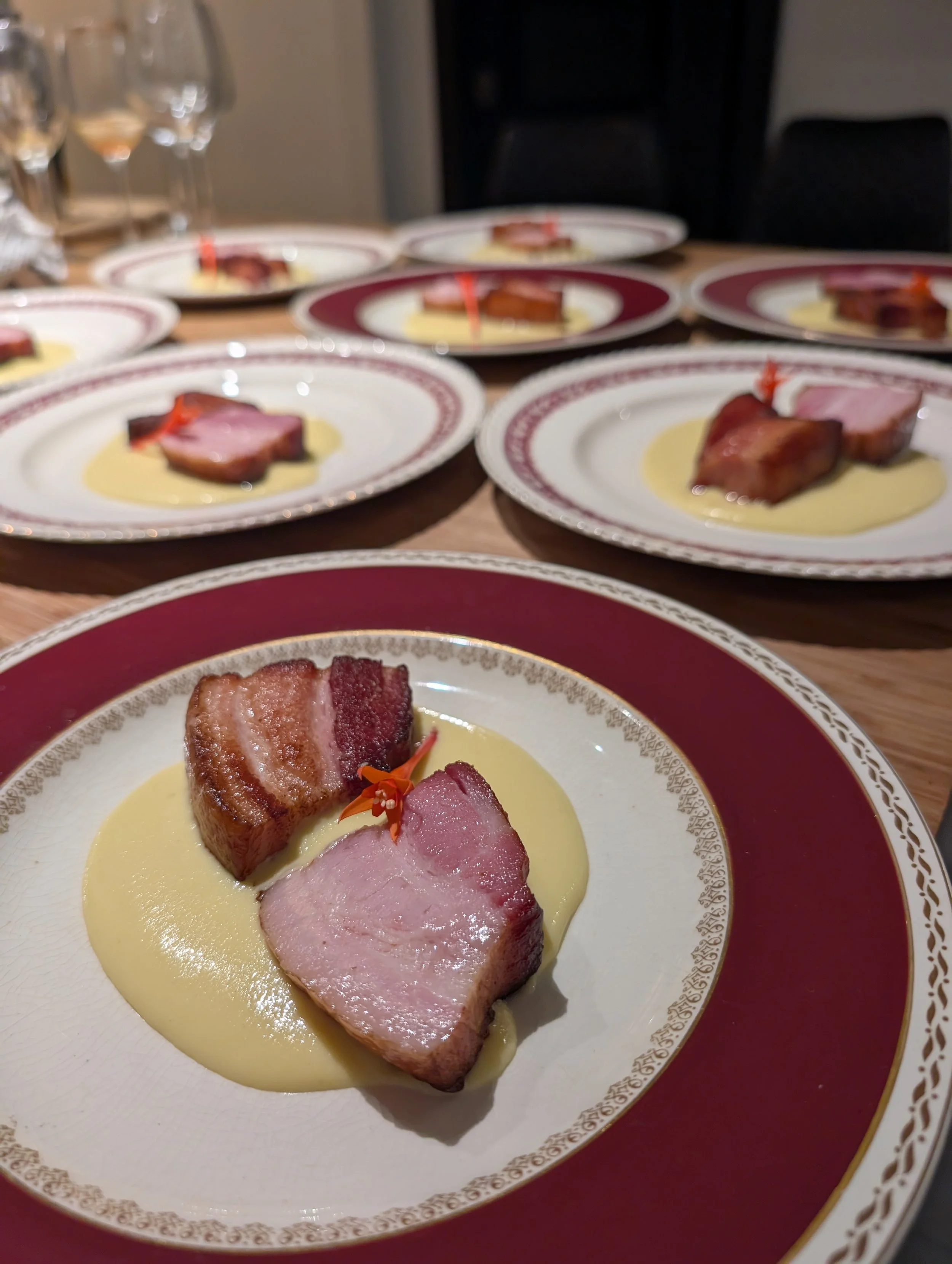 Assiette avec tranches de viande servies dans une sauce jaune, décorée d'une petite fleur orange, sur une assiette blanche avec bordures dorées et rouges, en arrière-plan plusieurs autres assiettes similaires, verres à vin en haut à gauche.