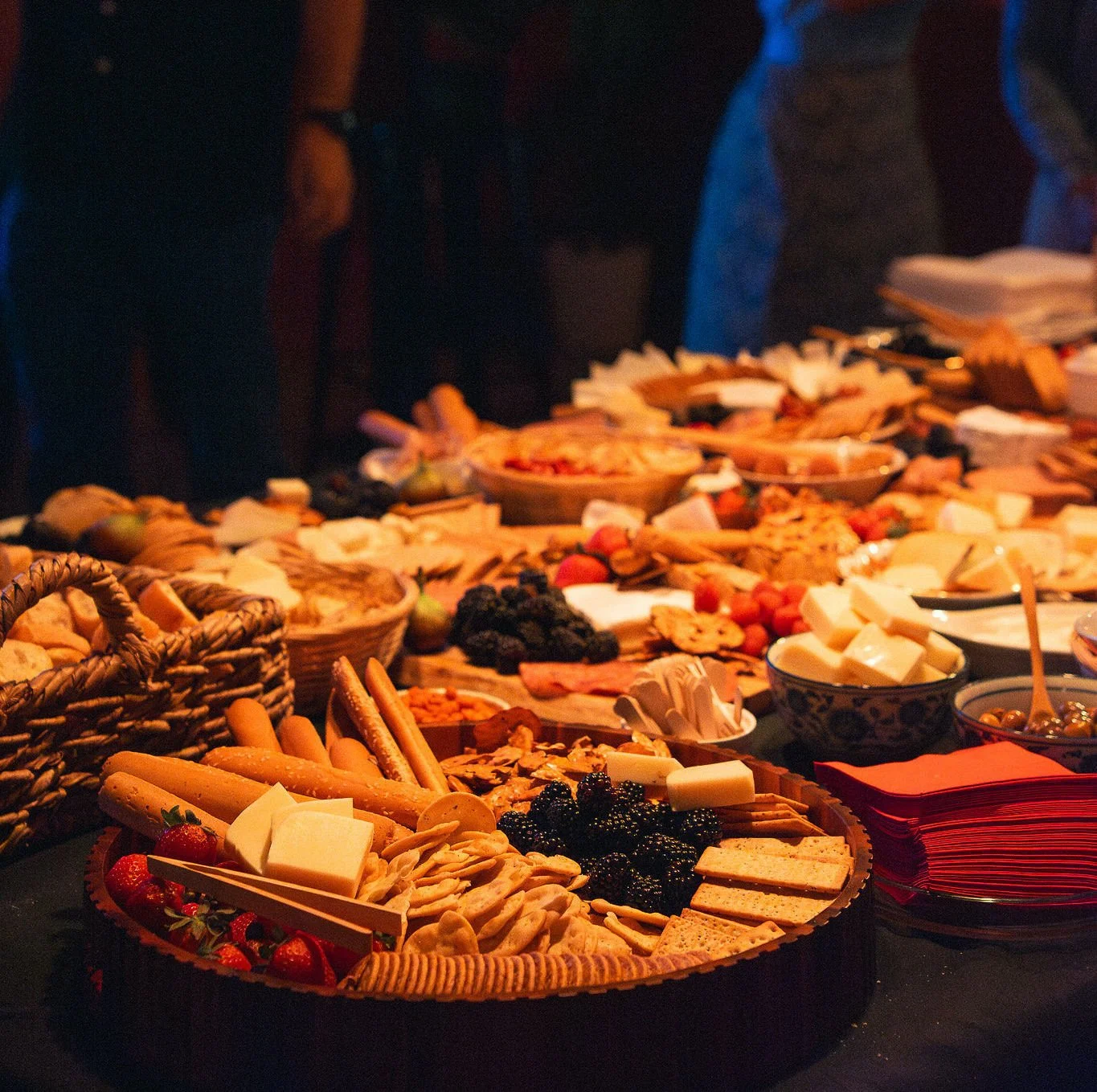 Table garnie d'un assortiment de fromages, biscuits, fruits, et autres amuse-bouches, avec plusieurs plats en arrière-plan dans une ambiance chaleureuse.