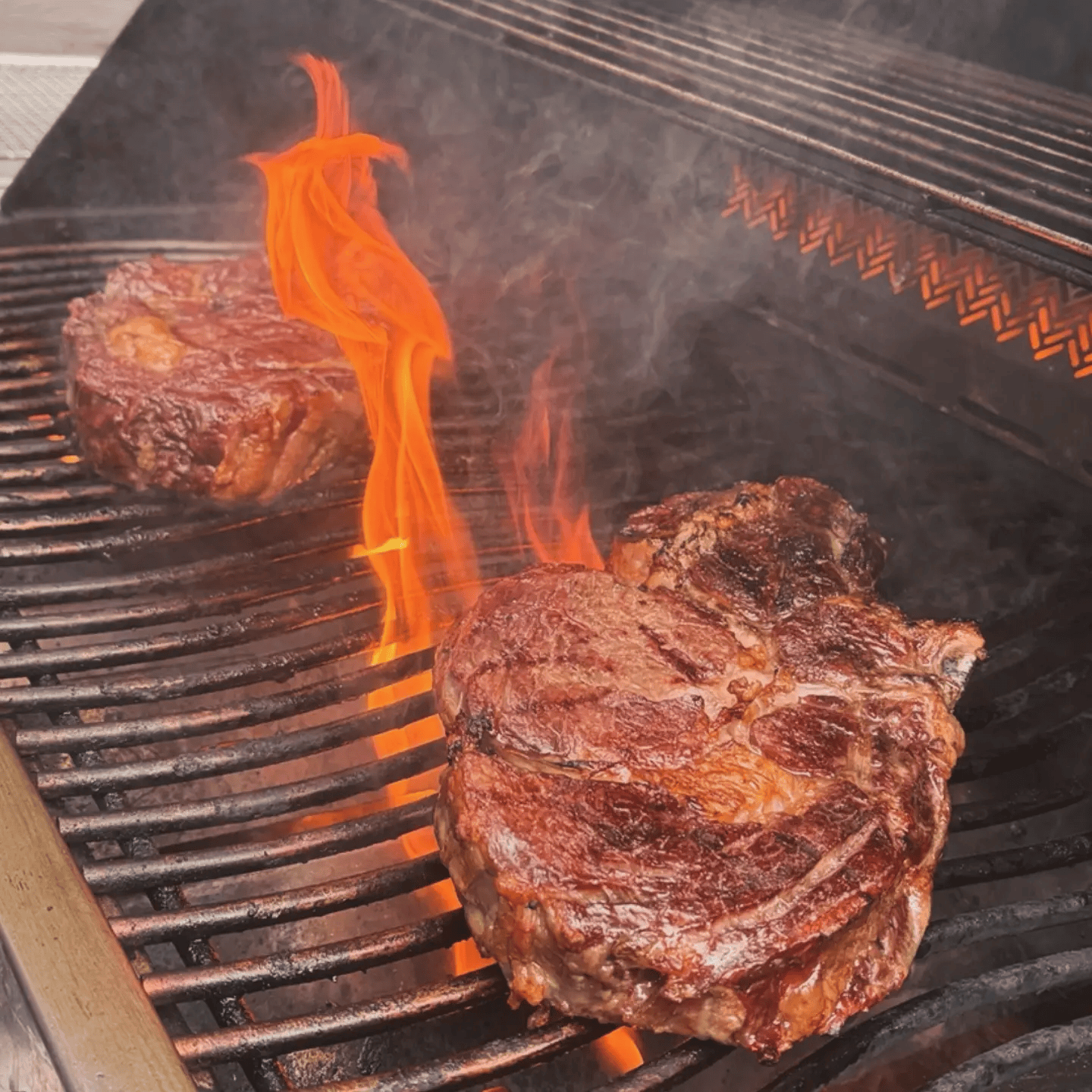 Deux morceaux de viande en train de griller sur un barbecue à charbon, avec des flammes oranges et de la fumée.