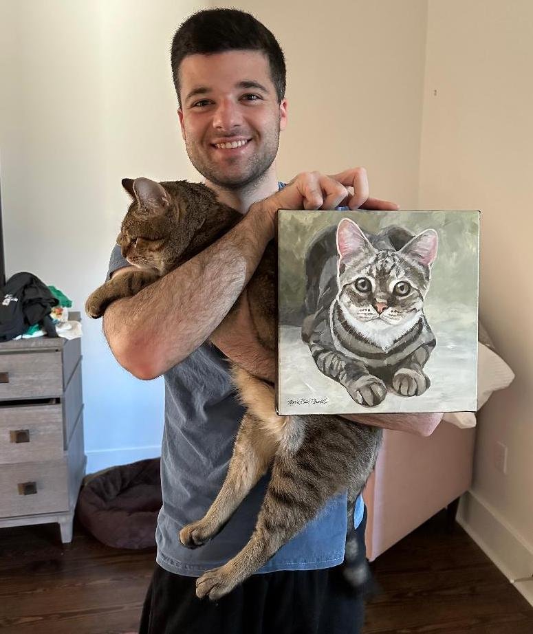 Photo of a smiling man holding his cat and a painting of his cat