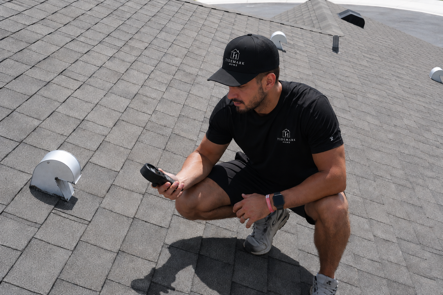 A man in black athletic clothing and a black cap squats on a rooftop, looking at his phone, with vents and roofing shingles visible.