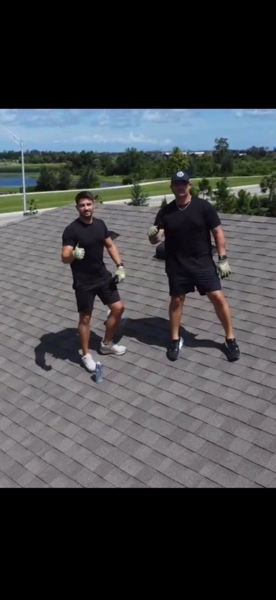 Two men wearing black shirts, shorts, and gloves standing on a rooftop, posing with fists up, with a scenic background of trees, water, and cloudy sky.