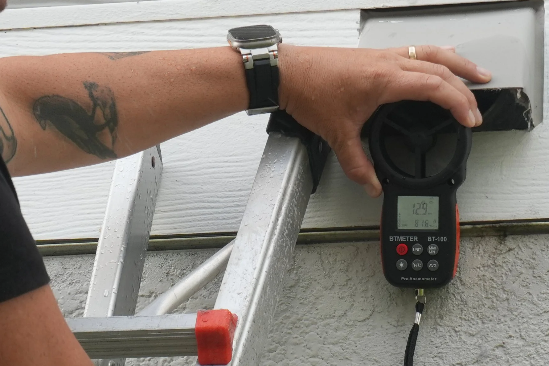A person's hand holding a digital anemometer measuring wind speed, with a ladder and a house exterior in the background.