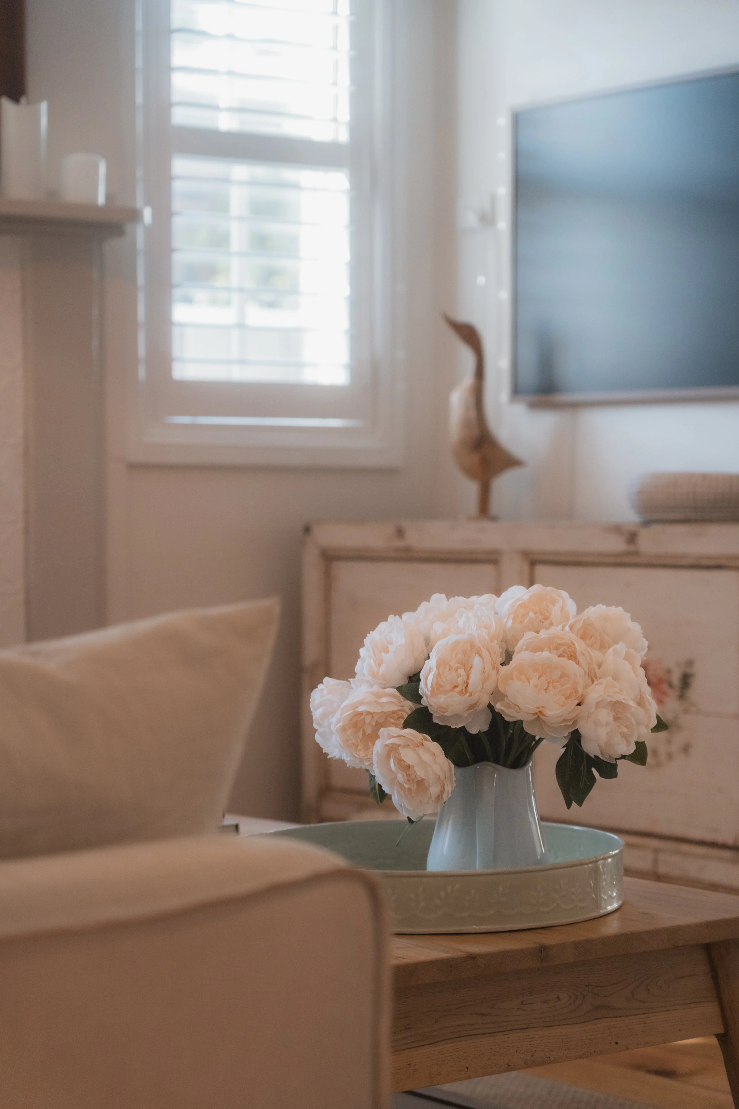 A white vase with a bouquet of peach-colored peonies on a round tray, placed on a wooden table in a bright room with a large window and shutters.