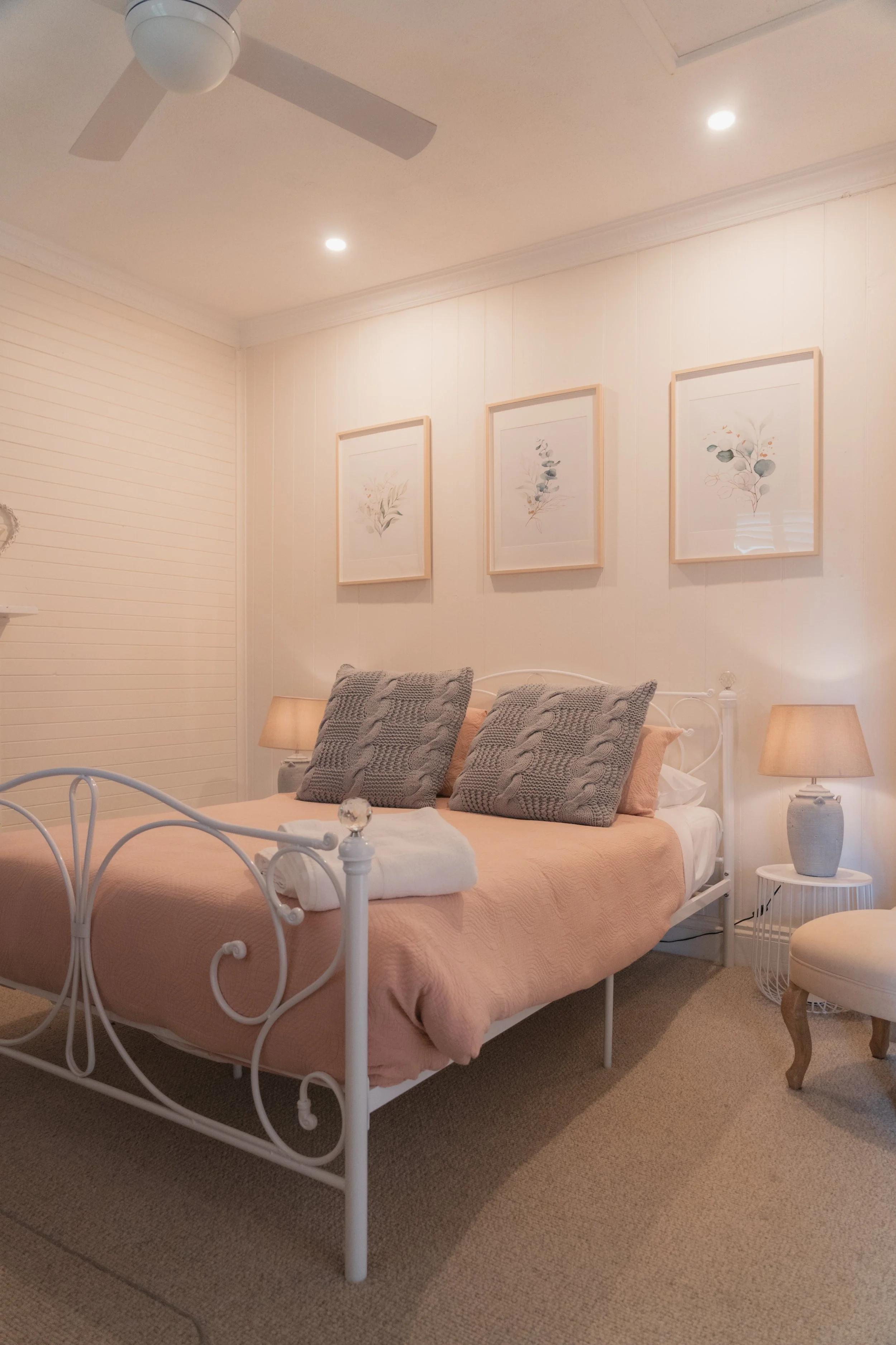 A cozy bedroom with a white metal bed frame, pink bedspread, and three large knitted gray pillows. On each side of the bed is a small wooden nightstand with a beige table lamp. The wall above the bed features three framed botanical watercolor prints.
