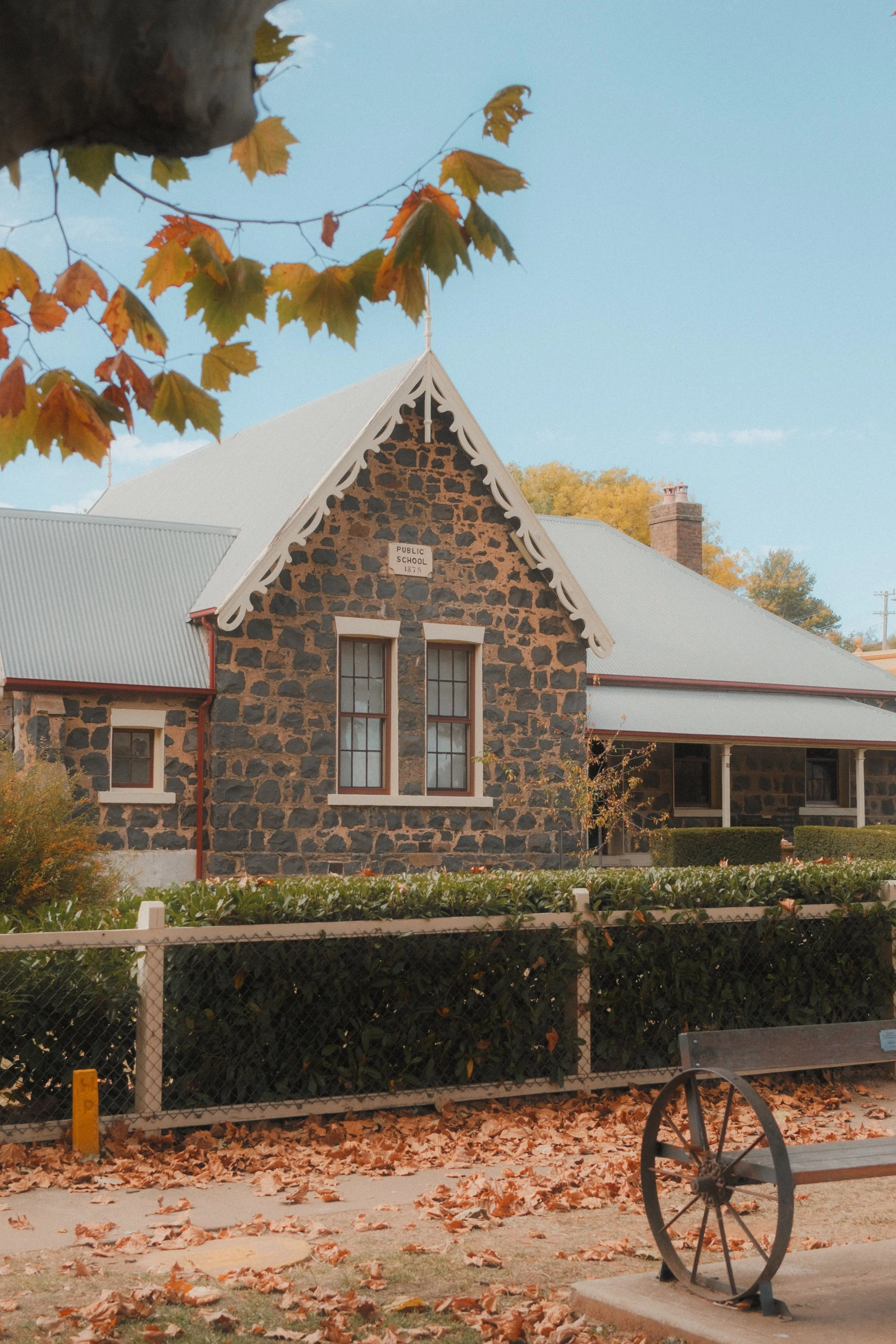 A historic stone schoolhouse with a sign that reads 'Public School 1875,' set behind a chain-link fence and surrounded by fallen autumn leaves, with a picnic bench and a large wheel in the foreground.