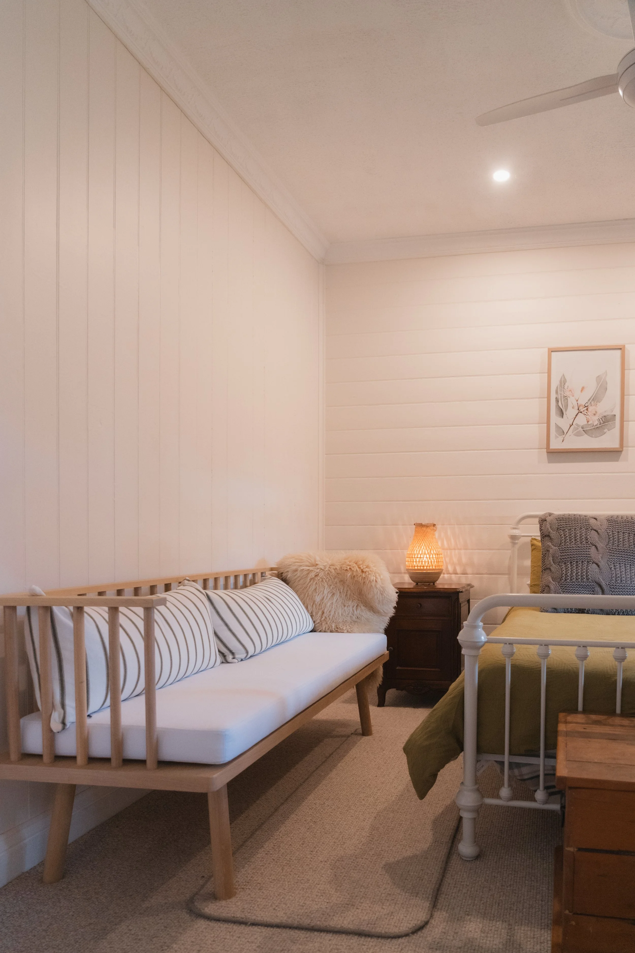 A cozy bedroom with a white wooden bed, green bedding, and a bedside table with a lamp. The wall is decorated with a framed botanical print, and there is a ceiling fan and recessed lighting. A gray armchair with a knit blanket is visible on the right