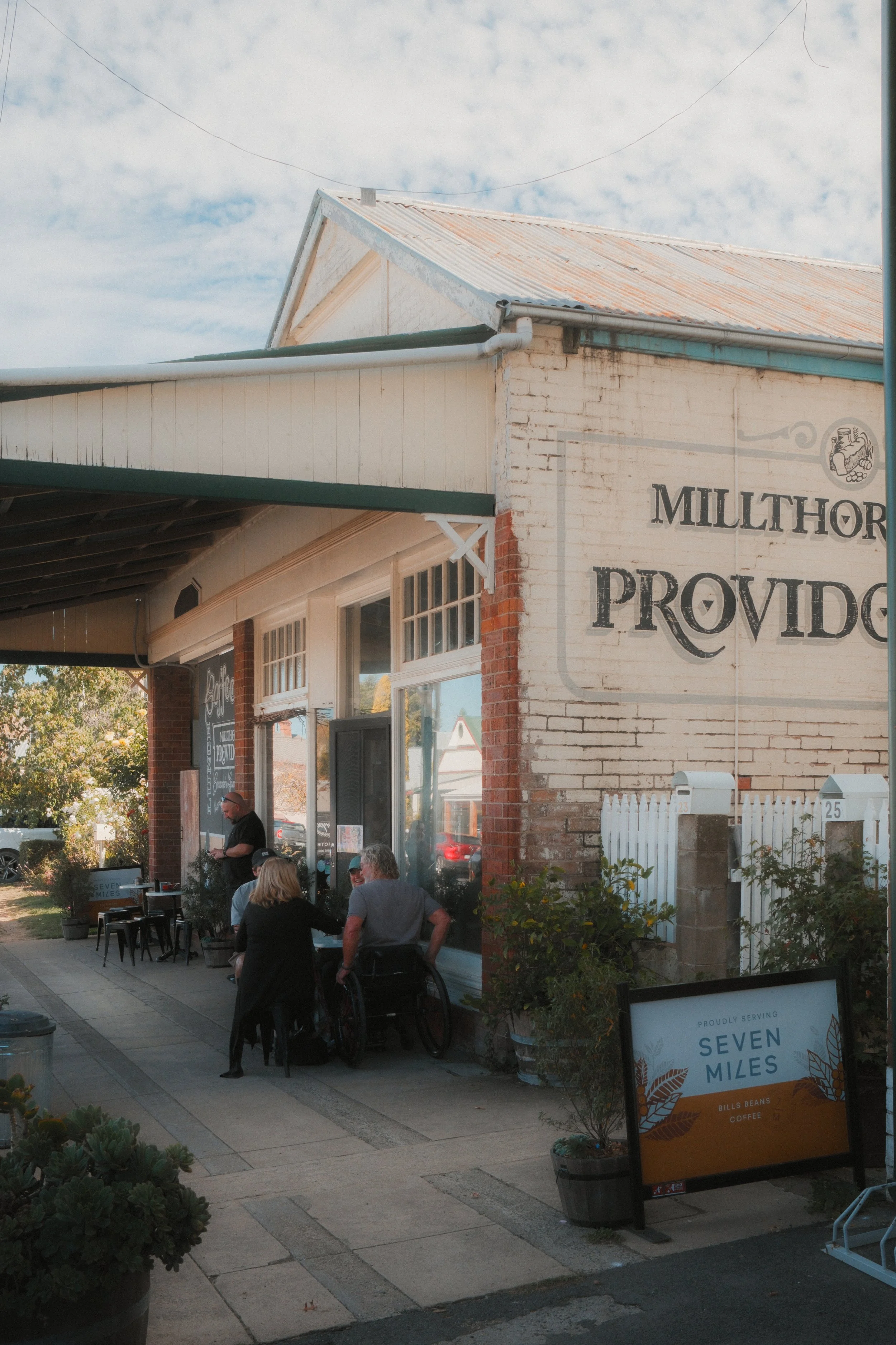 Exterior of a café named 'Seven Miles' with outdoor seating, people gathered and conversing, a signboard displaying the name and coffee offerings, and a brick building with a white and brick facade under a partly cloudy sky.