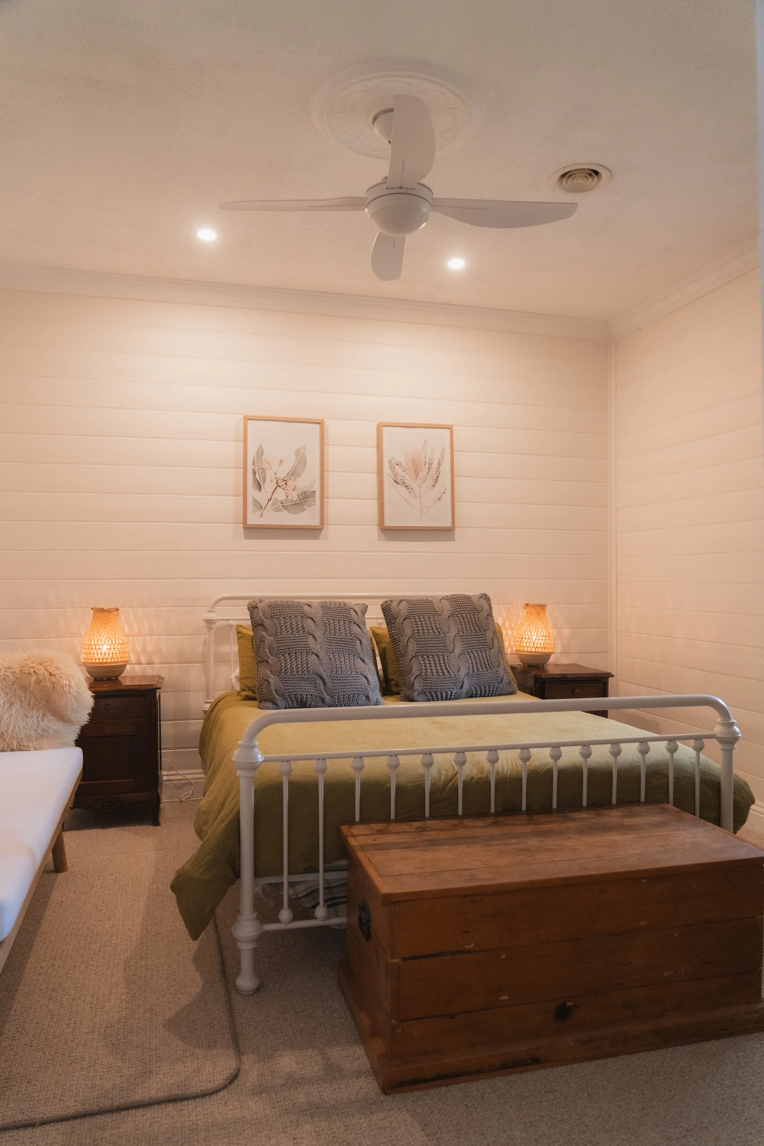 A bedroom with a white metal bed frame, green bedding, and three gray patterned pillows. Two wooden nightstands with wicker lamps on either side of the bed. Two framed botanical prints on the beige paneled wall. A wooden chest at the foot of the bed.