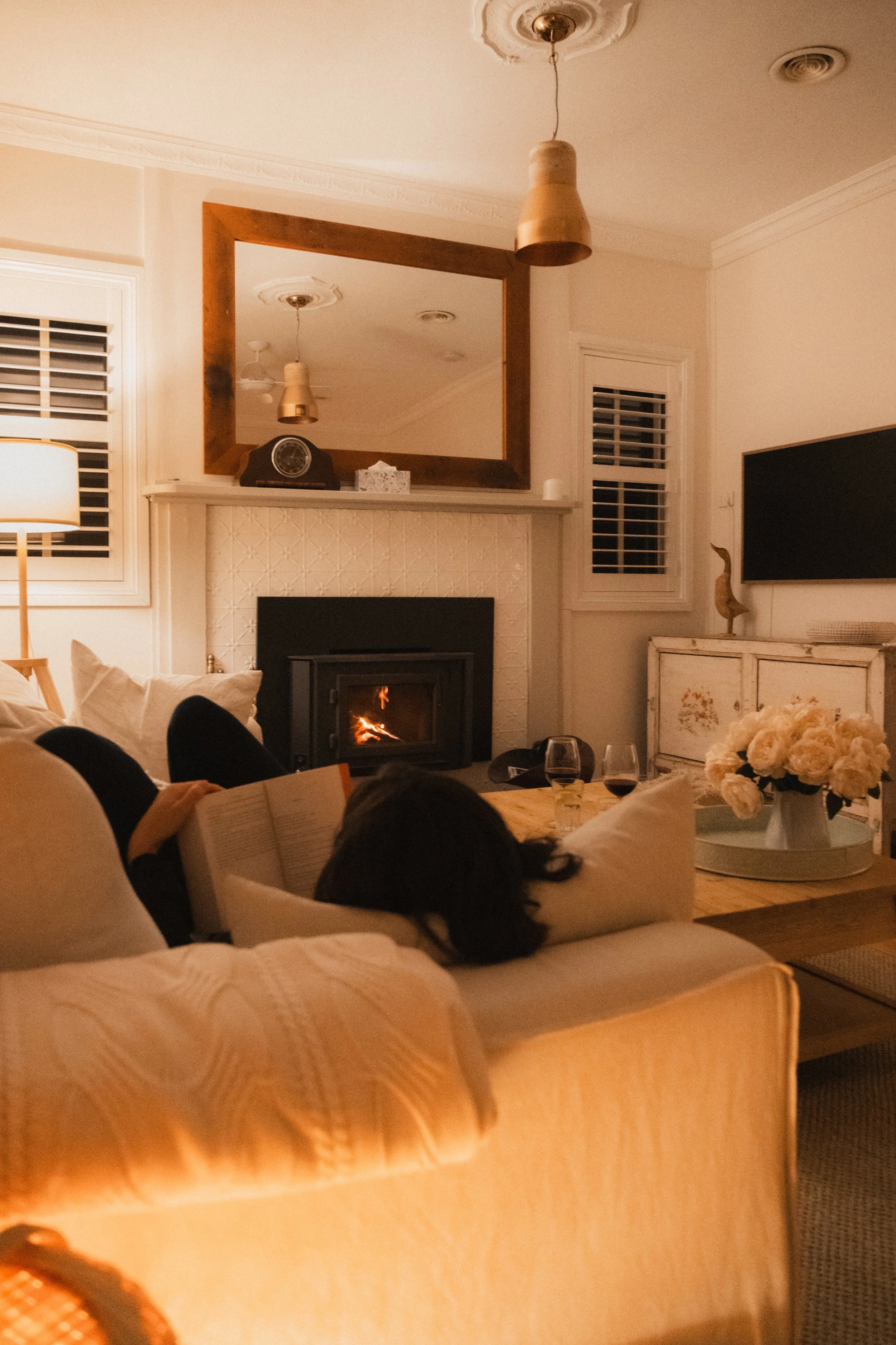 Warm living room at Church Cottage Millthorpe with a lit fireplace, soft sofa, wine glasses on the coffee table and a guest reading by the fire.