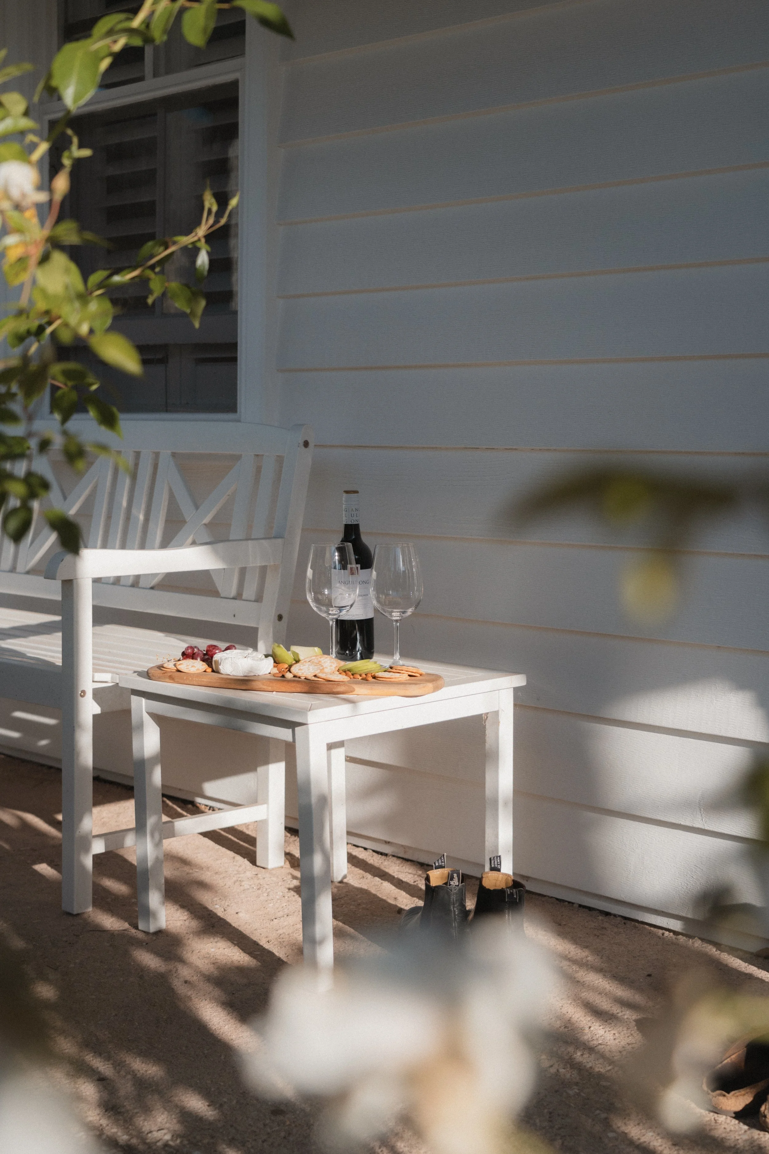 Outdoor scene with a white wooden bench and table set against a light-colored house wall, with a bottle of wine and two wine glasses, cheese and crackers on the table, and shoes on the ground, partially obscured by blurred foliage in the foreground.