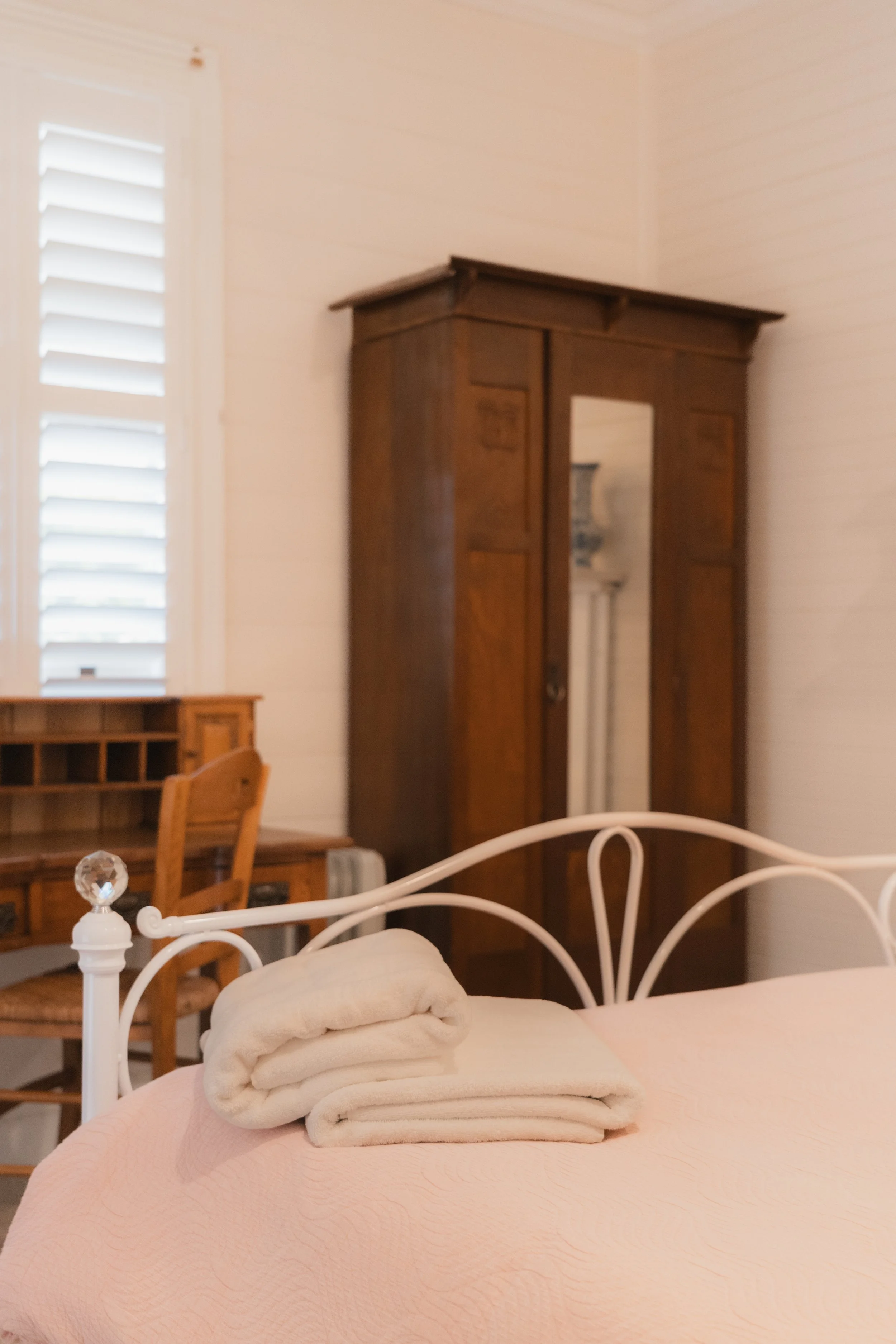 A bed with pink bedding and rolled towels on top, in a cozy bedroom with wooden furniture, including a dresser and a tall cabinet, and a window with white shutters.