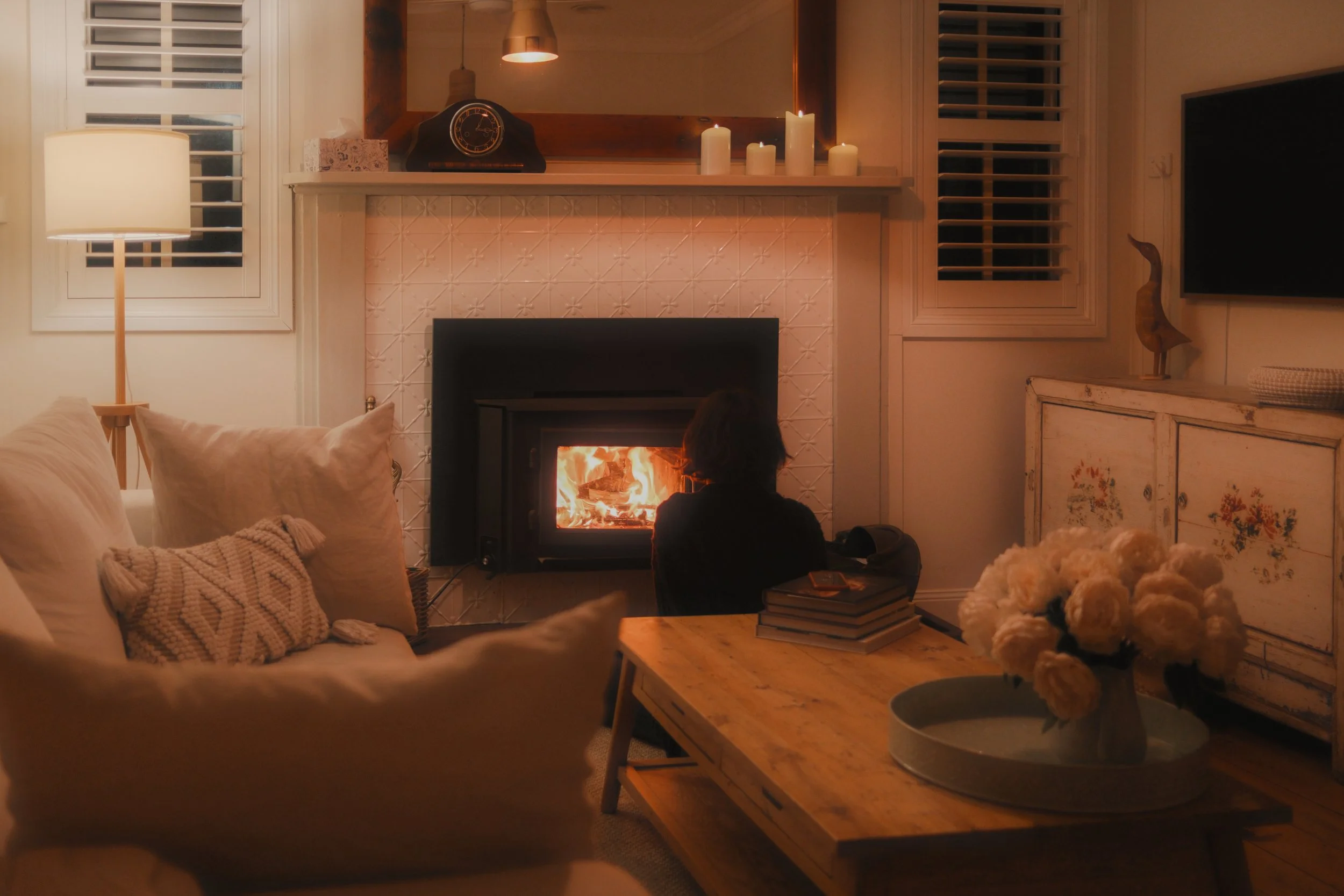 A cozy living room with a lit fireplace, candles on the mantel, a person sitting on the floor facing the fire, a beige sofa with pillows, a wooden coffee table with a stack of books, and a vase of white flowers on a side table.