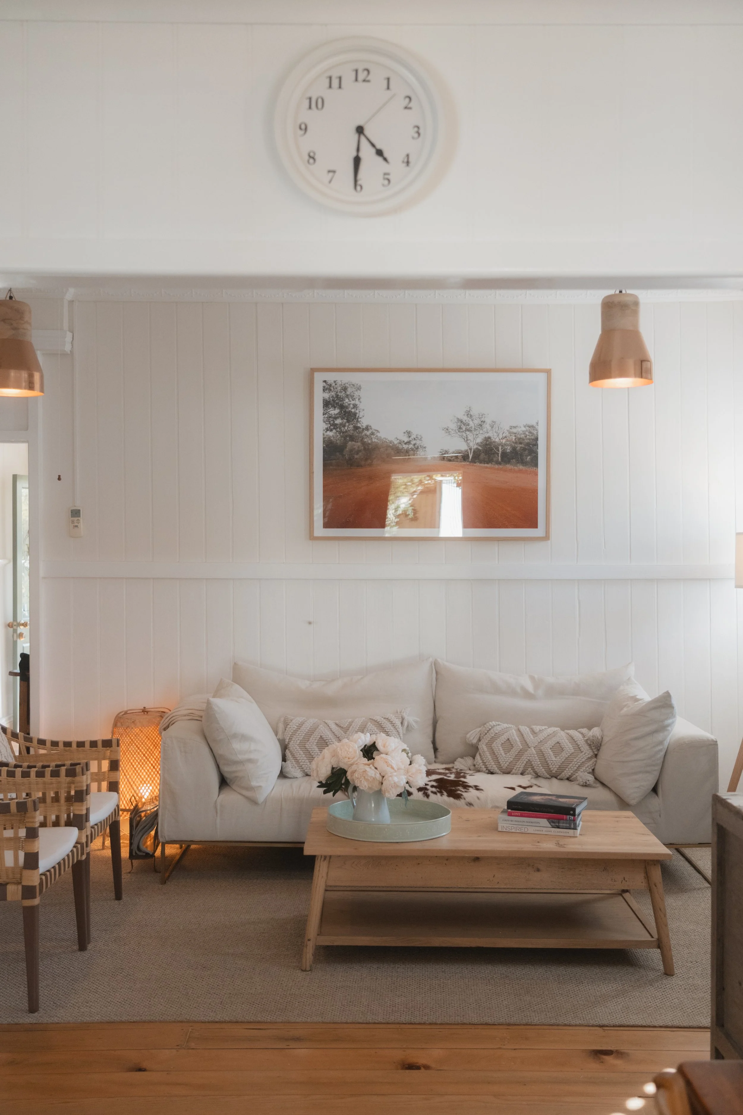 A cozy living room with a white sofa decorated with patterned pillows, a wooden coffee table holding a vase of white flowers and books, surrounded by woven chairs, pendant lights, a framed landscape photo on the wall, and a wall clock showing 6:08.