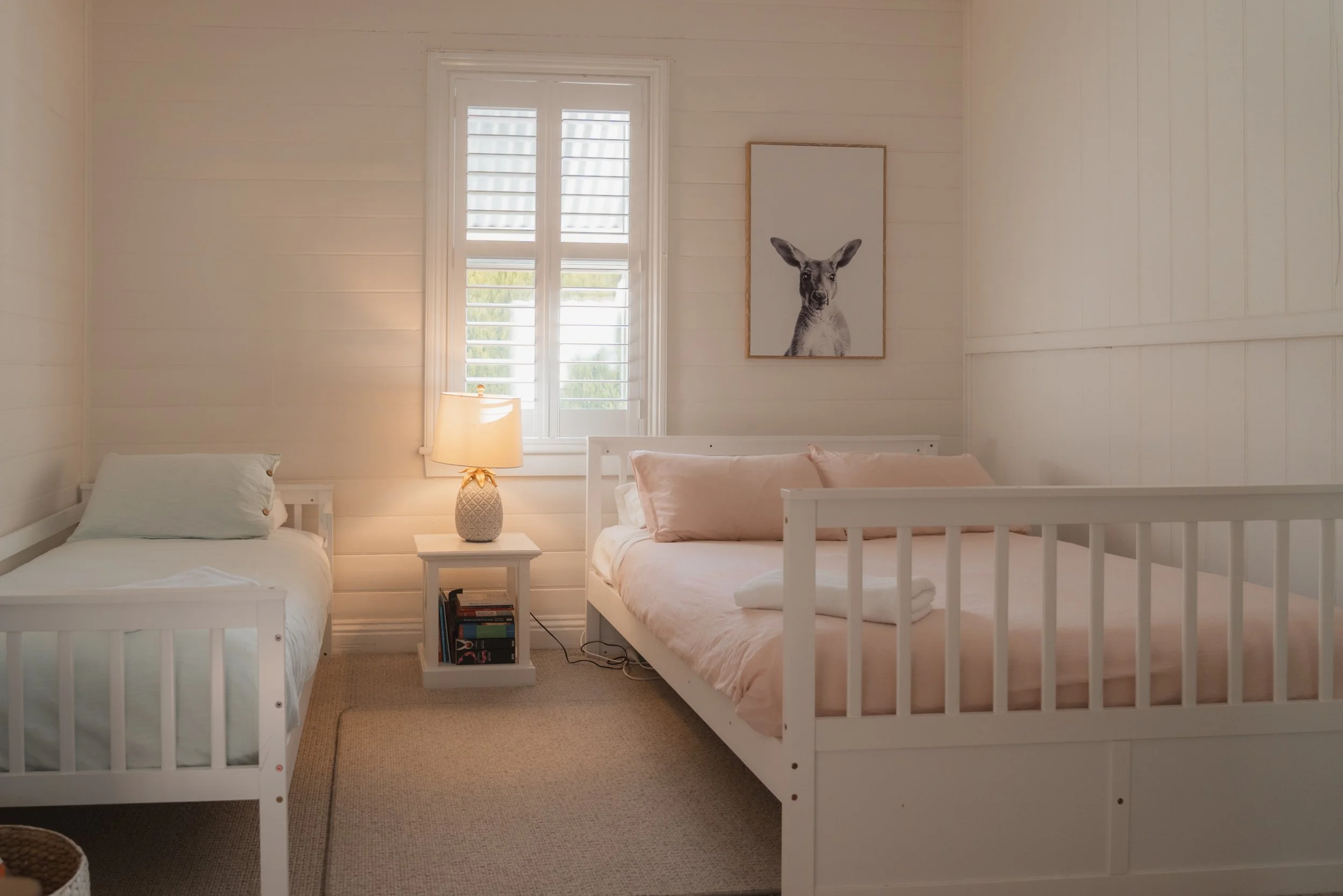 A bedroom with two white twin beds, a window with white shutters, a nightstand with a lamp and books, and a framed drawing of a deer on the wall.